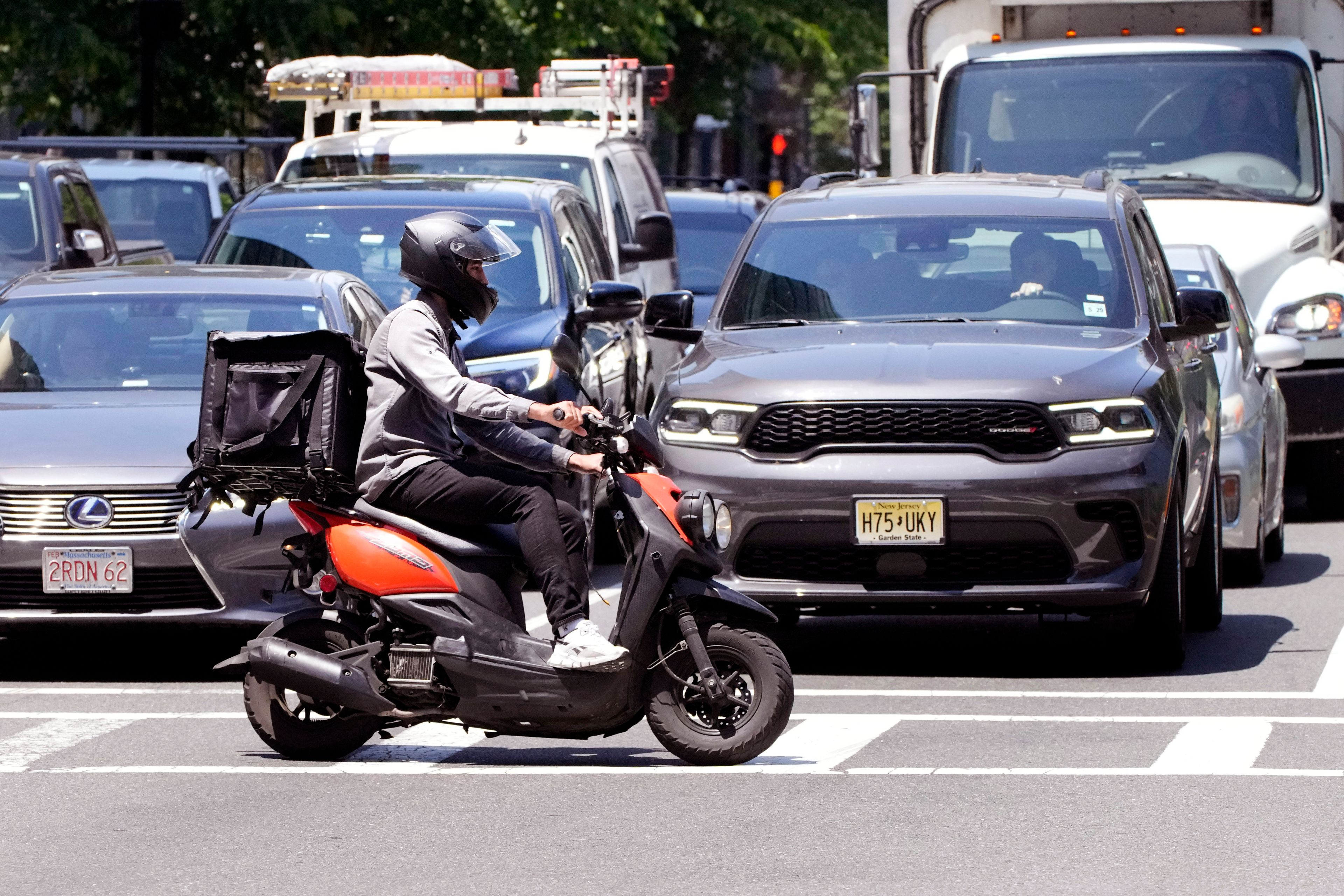 A delivery driver on a scooter rides the pedestrian crosswalk through traffic on a delivery in the Seaport District, Friday, June 7, 2024, in Boston. A soaring demand for food delivered fast has spawned small armies of couriers in a growing number of cities where delivery scooters, motorcycles and mopeds zip in and out of traffic and hop onto sidewalks alongside startled pedestrians racing to drop off salads and sandwiches.