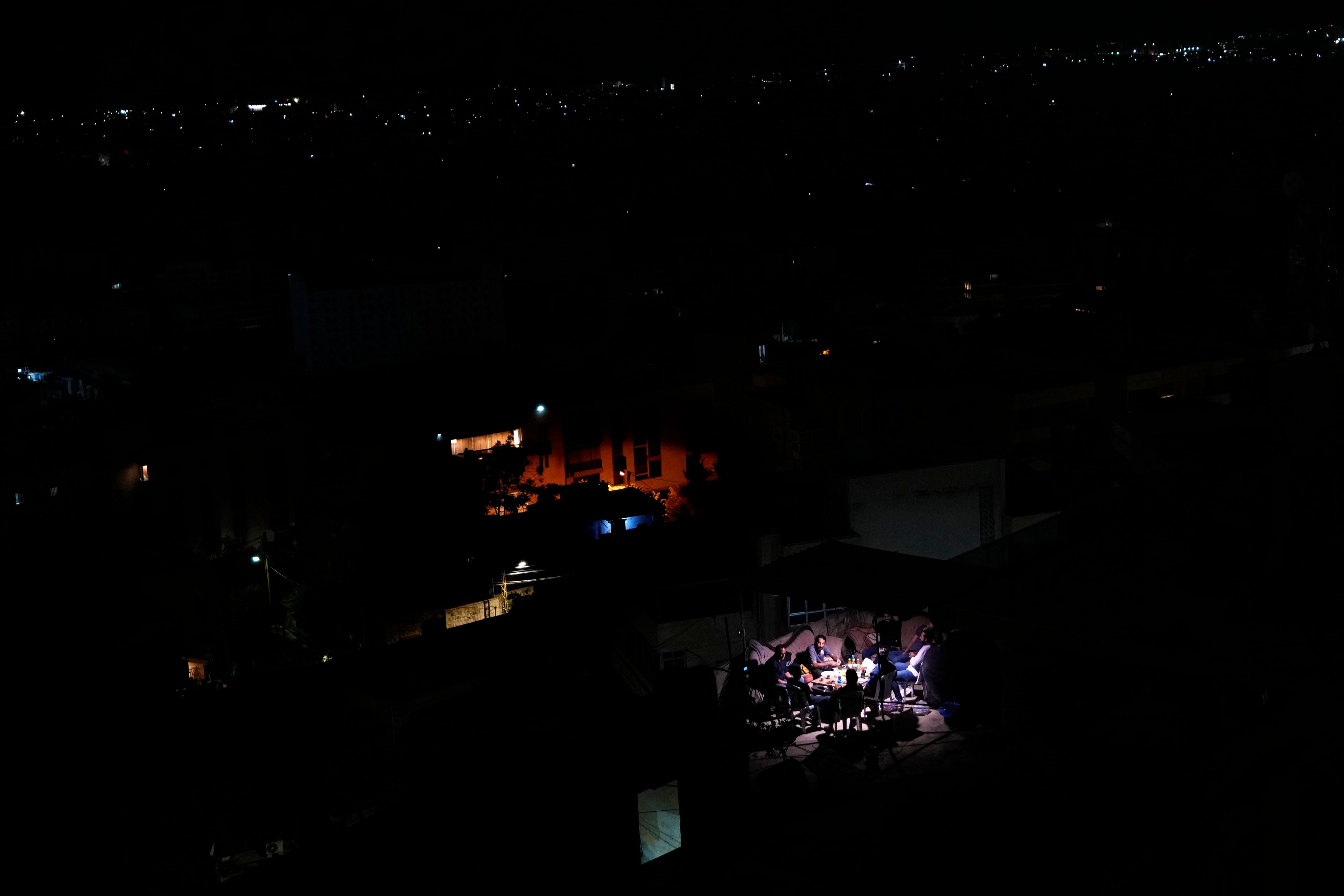 Residents sit on the roof of a building and have dinner as Dahiyeh suburb, background, remains in darkness after Israeli airstrikes, Lebanon, Friday, Oct. 11, 2024. (AP Photo/Hassan Ammar)