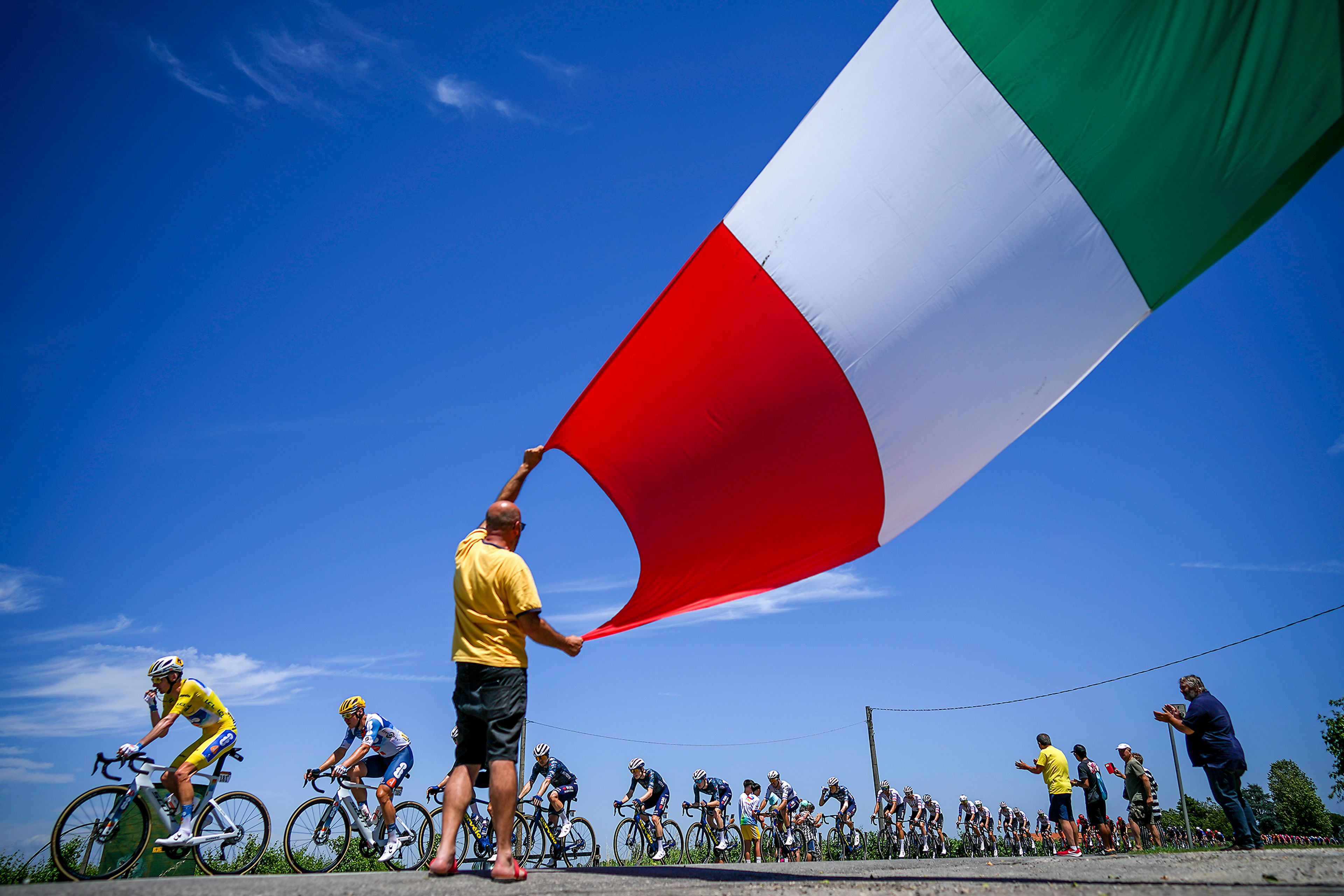 A cycling fan waves the Italian flag as the peloton with Team dsm–firmenich PostNL’s Romain Bardet, of France, wearing the yellow jersey, left, passes during the second stage of the Tour de France over 199.2 kilometers with start in Cesenatico and finish in Bologna, Italy, Sunday, June 30, 2024. It was the first time in the race’s 121-year history, that it has started in Italy. 2024 is the 100th anniversary of the first Italian victory in the Tour, won by Ottavio Bottecchia in 1924. (AP Photo/Daniel Cole)