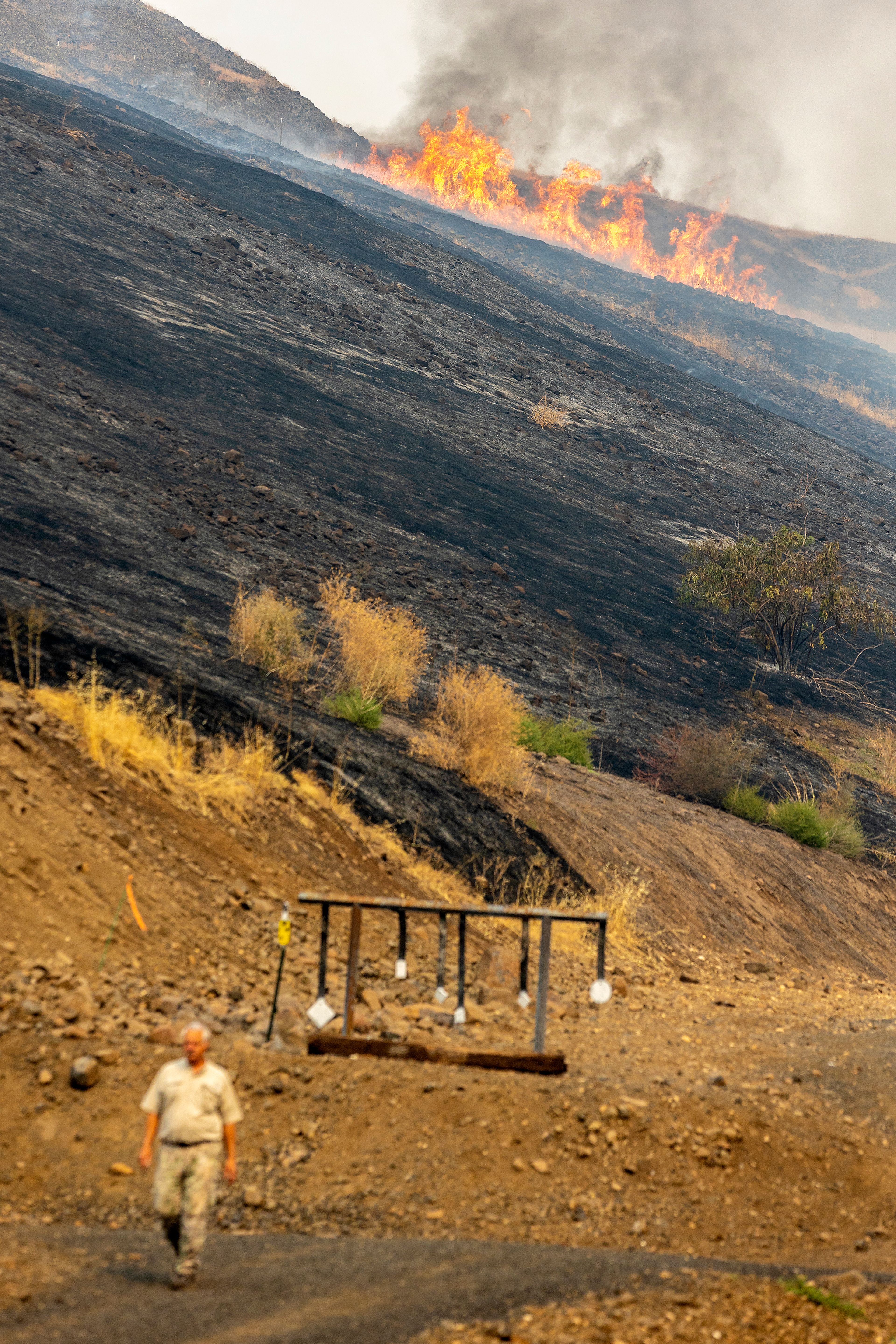 A man walks down a path Tuesday as flames burn along the hillside in the background at the Lewis-Clark Wildlife Club shooting range off South Tom Beall Road near Lapwai.