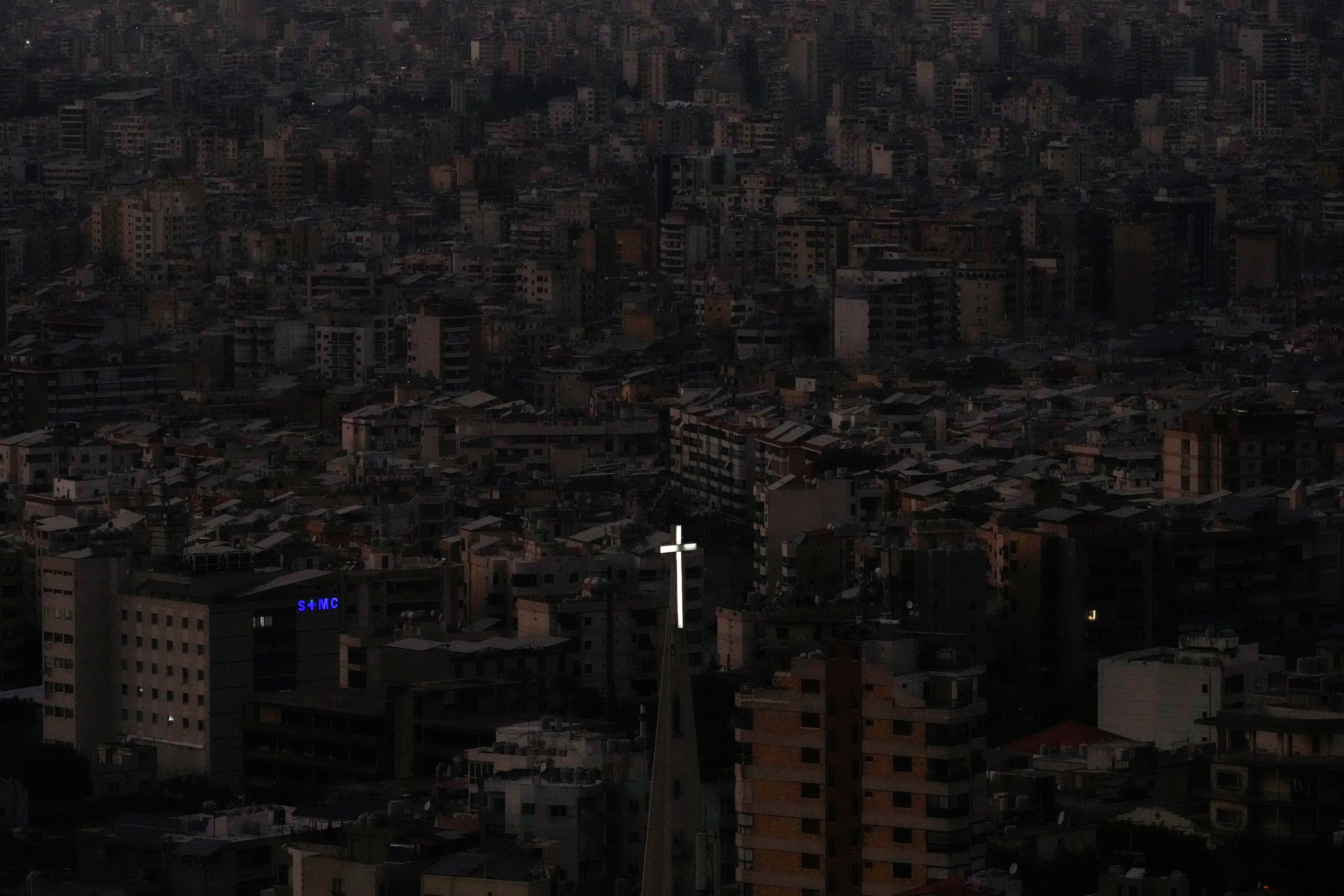 The cross atop of Our Lady of Hadath Church appears in front of Beirut's Dahiyeh suburb, background, that remains in darkness after Israeli airstrikes, Lebanon, Wednesday, Oct. 16, 2024. (AP Photo/Hassan Ammar)