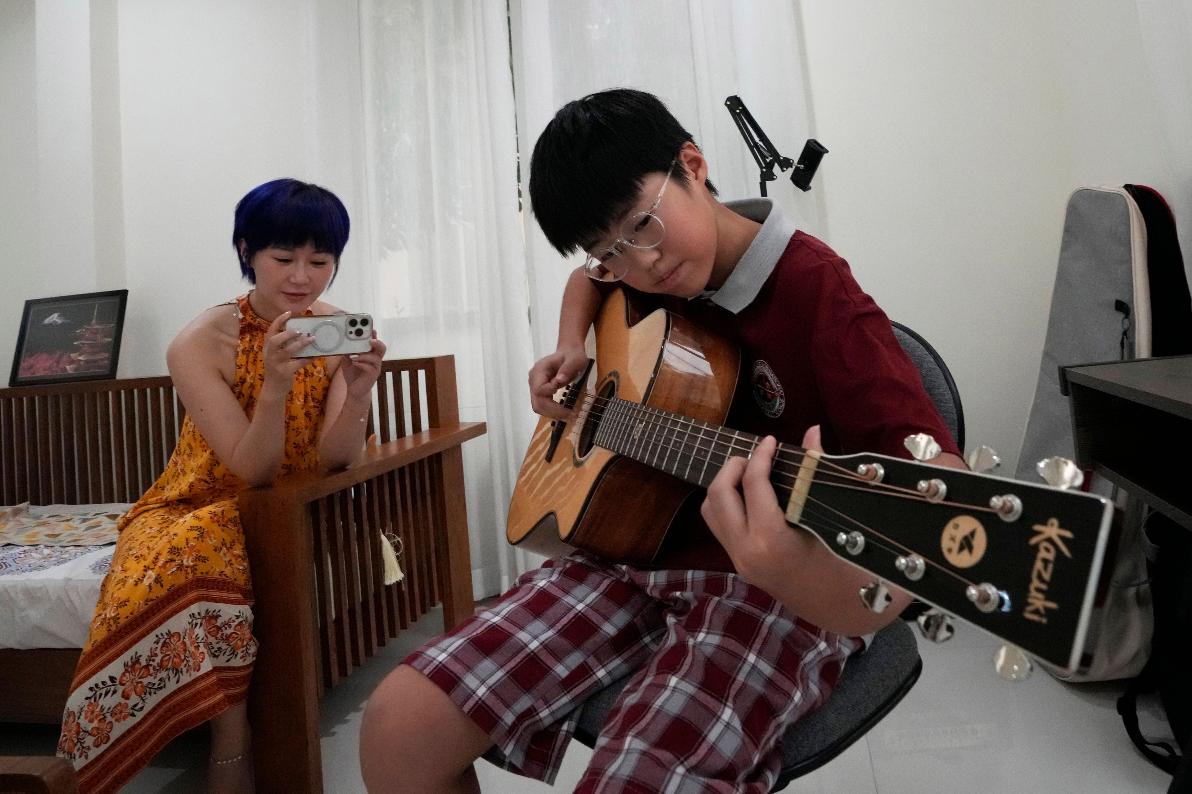 Chinese mother Jiang Wenhui, left, records her son Rodney Feng playing the acoustic guitar in Chiang Mai province, Thailand, Tuesday, April 23, 2024.