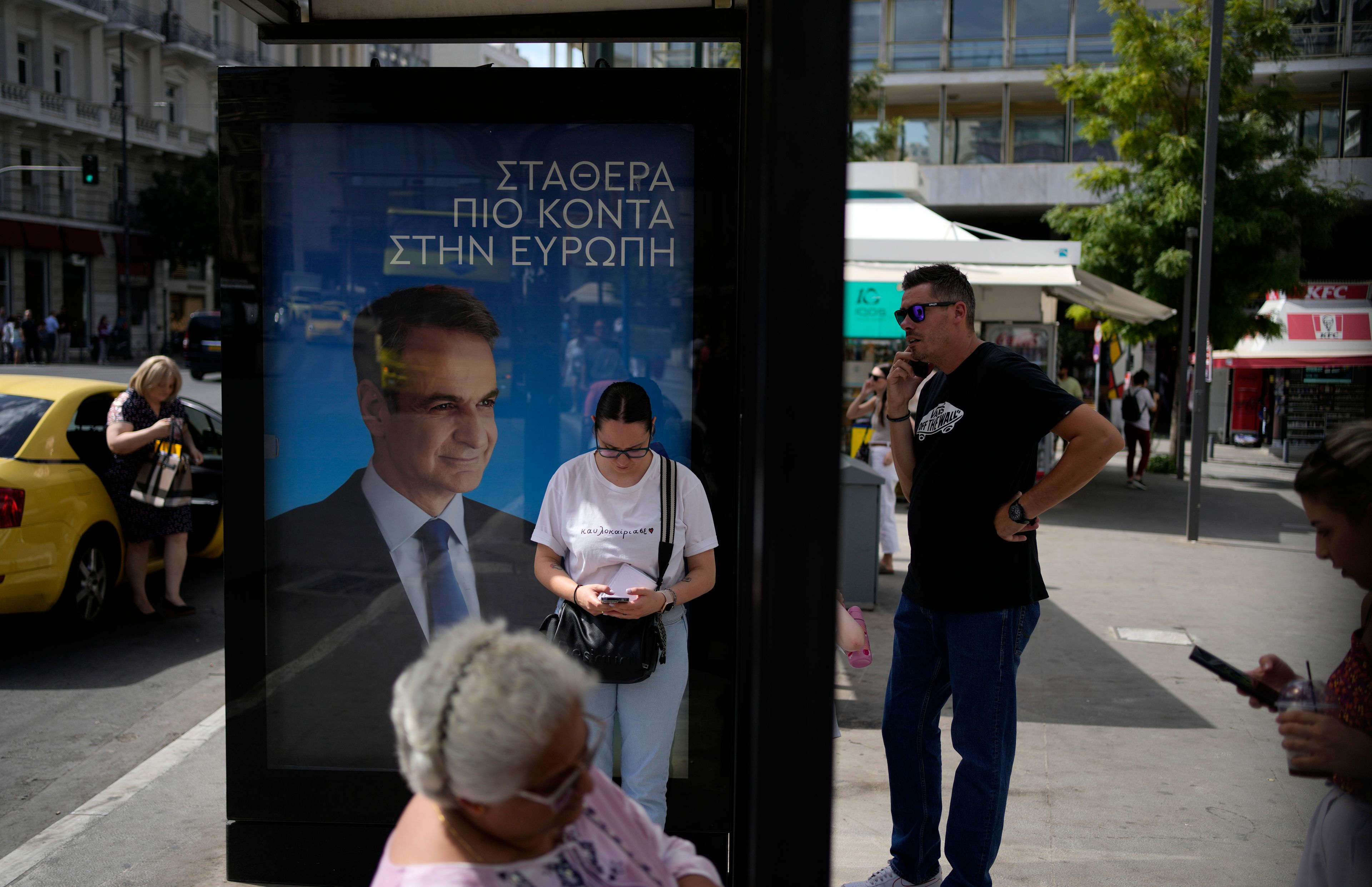 Commuters wait at a bus stop as an election poster depicting Greece's Kyriakos Mitsotakis reads "Steadily closer to Europe" in Athens, Greece, Friday, June 7, 2024. The European Election will take place on June 9.