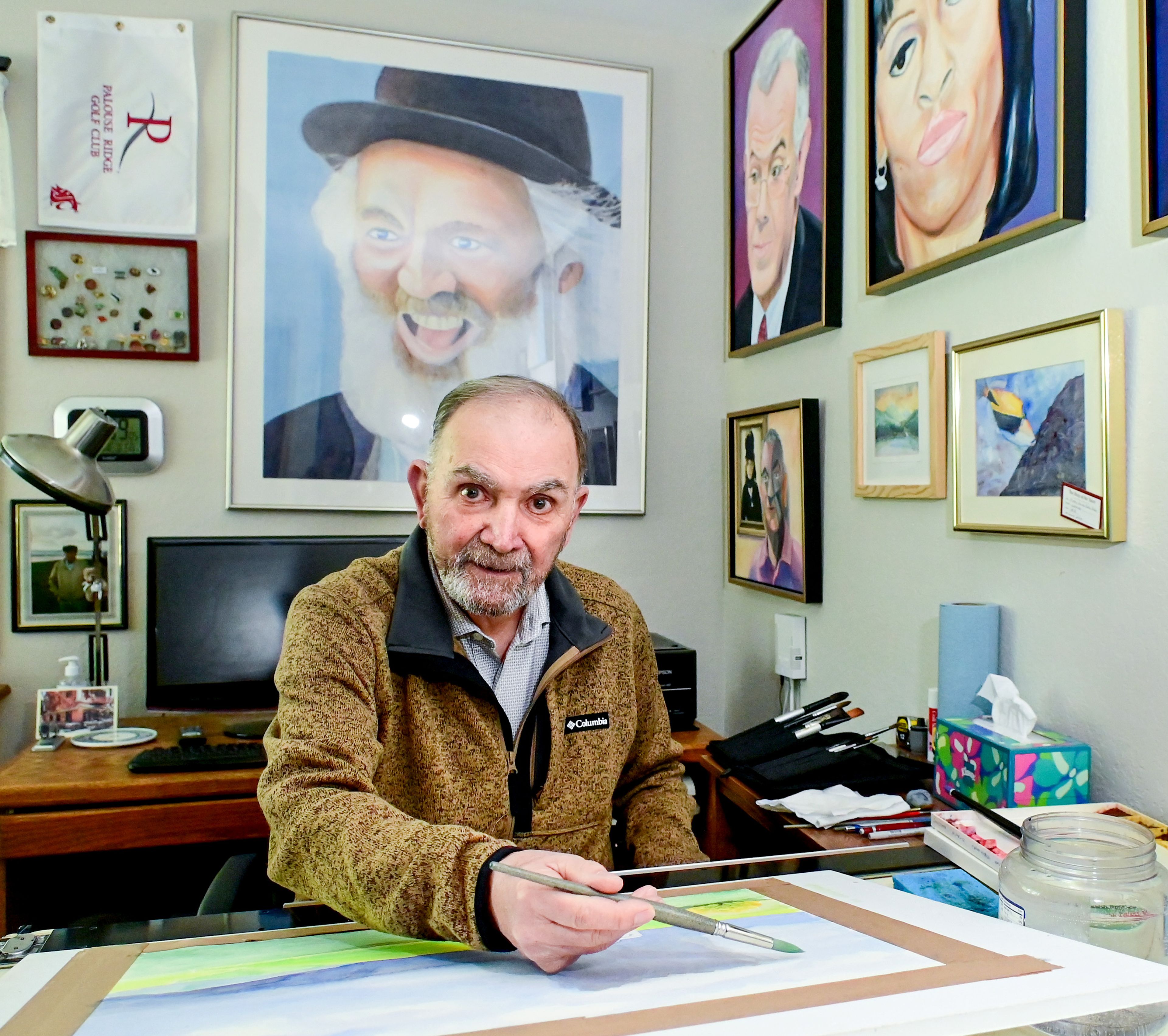 Ernie Weiss, president of the Palouse Watercolor Socius, sits in front of an in-progress watercolor piece, named “Abandoned,” that will be entered in the 50th anniversary show for the group. Other artwork by Weiss hangs on the walls around him.