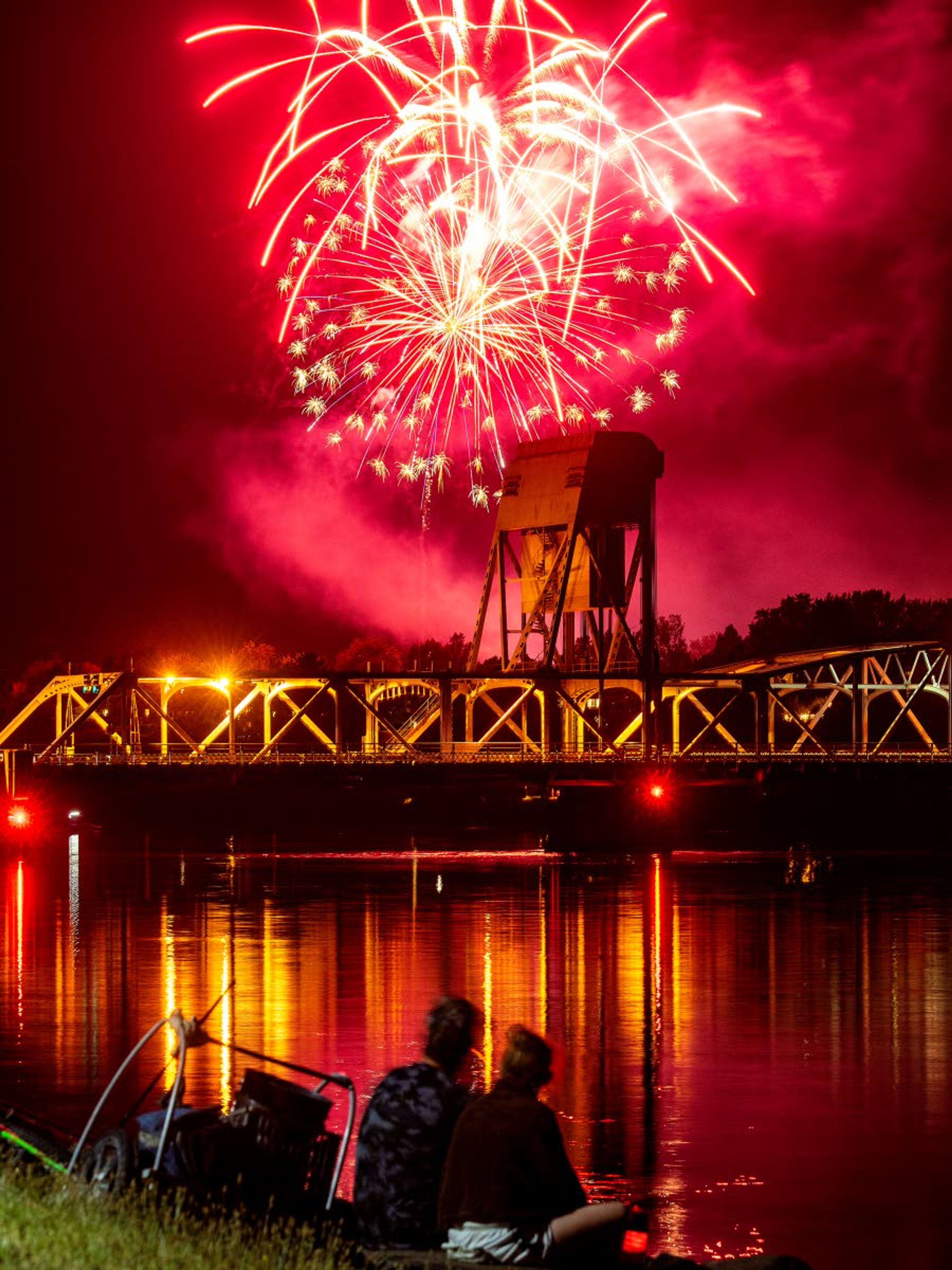 The Thursday evening air turns a bright red while fireworks explode over Clarkston as people sit along the Lewiston Levee Parkway Trail to watch the Community Spirit 34th Annual Fireworks Show 2019 from across the Snake River.