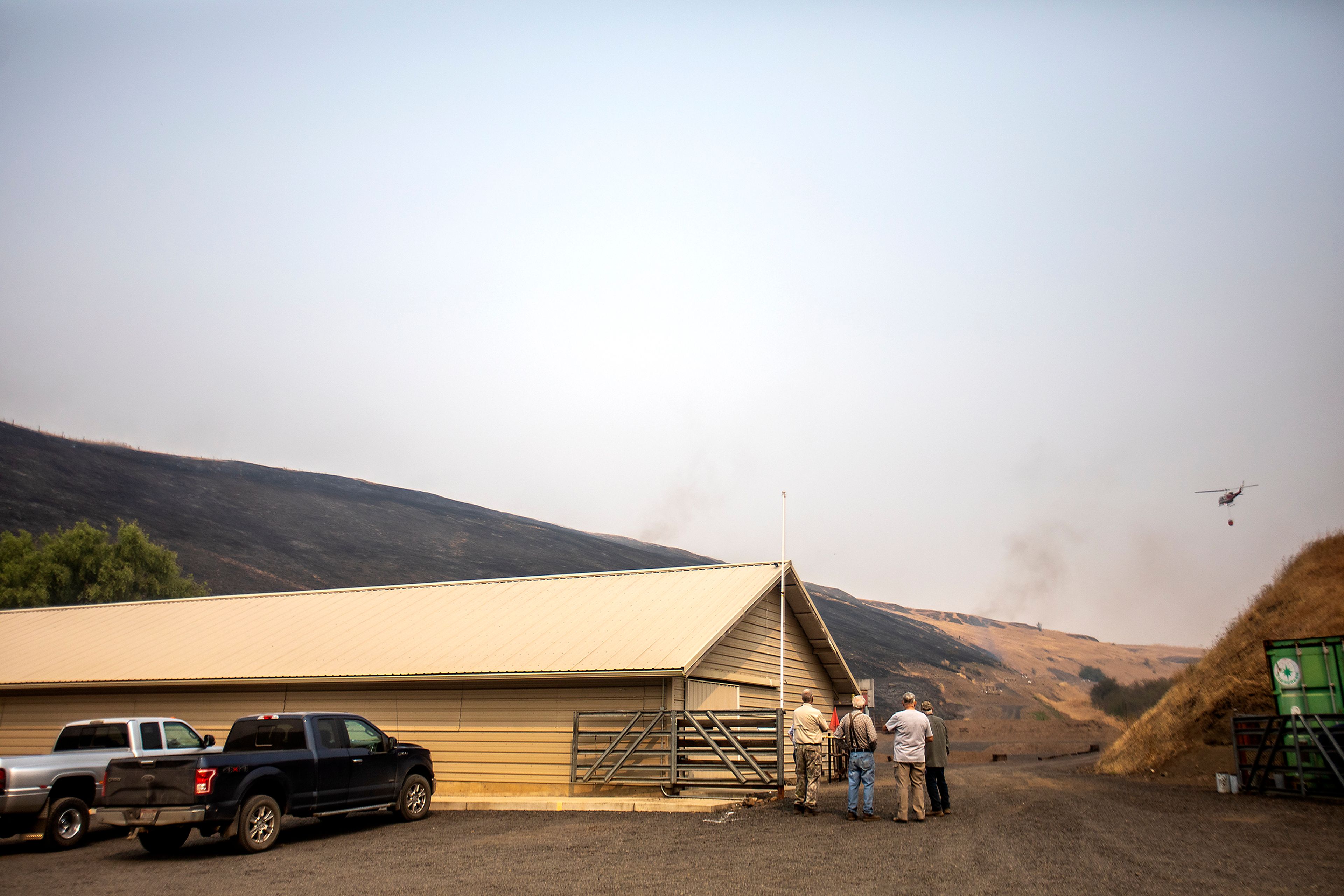 People stands outside the Lewis-Clark Wildlife Club shooting range Tuesday off South Tom Beall Road near Lapwai as a helicopter heads toward some flames to make a water drop.