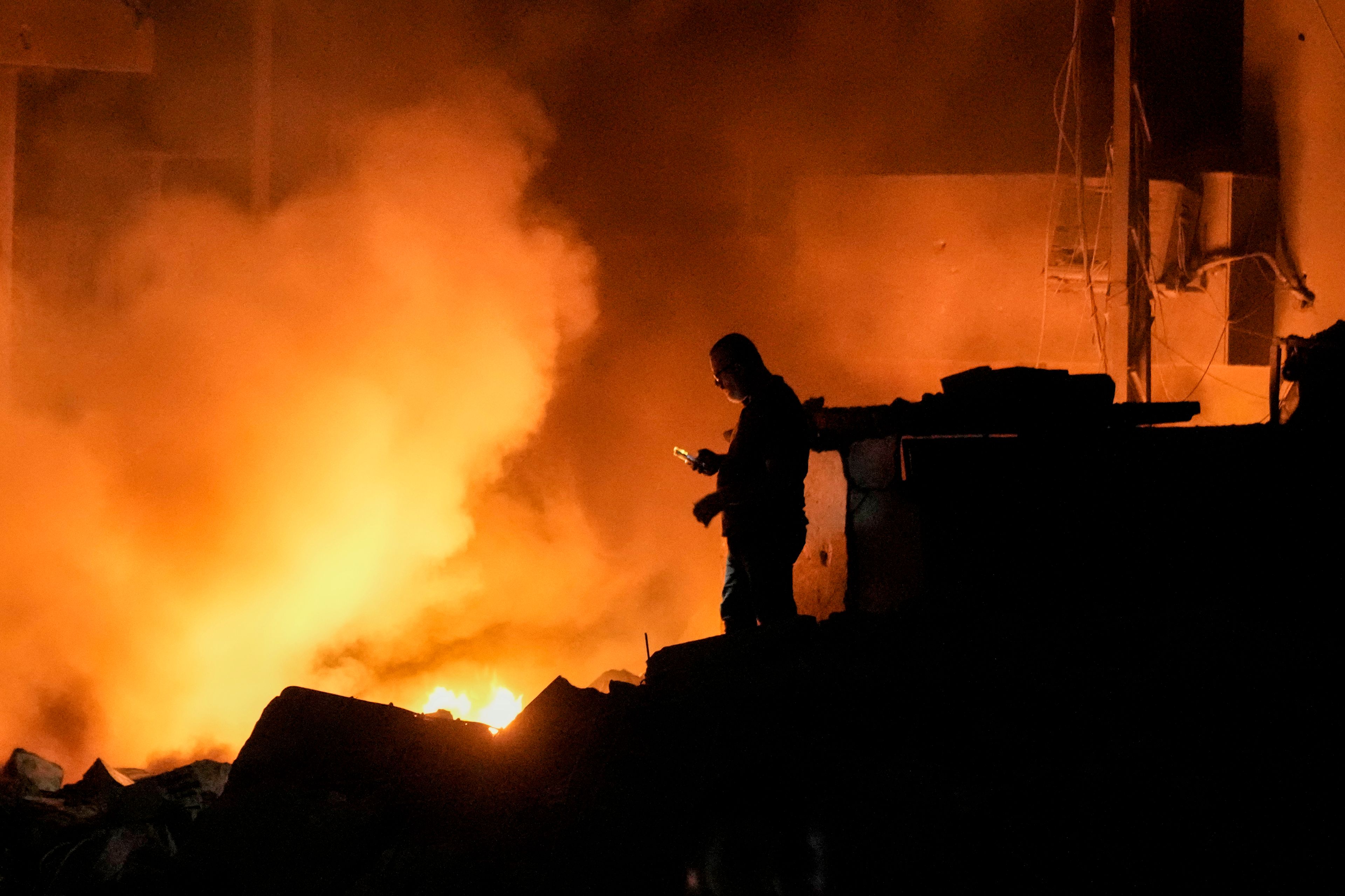 A man uses his mobile phone as flames and smoke rise at the scene of buildings hit by an Israeli airstrike in central Beirut, Lebanon, Thursday, Oct. 10, 2024. (AP Photo/Bilal Hussein)