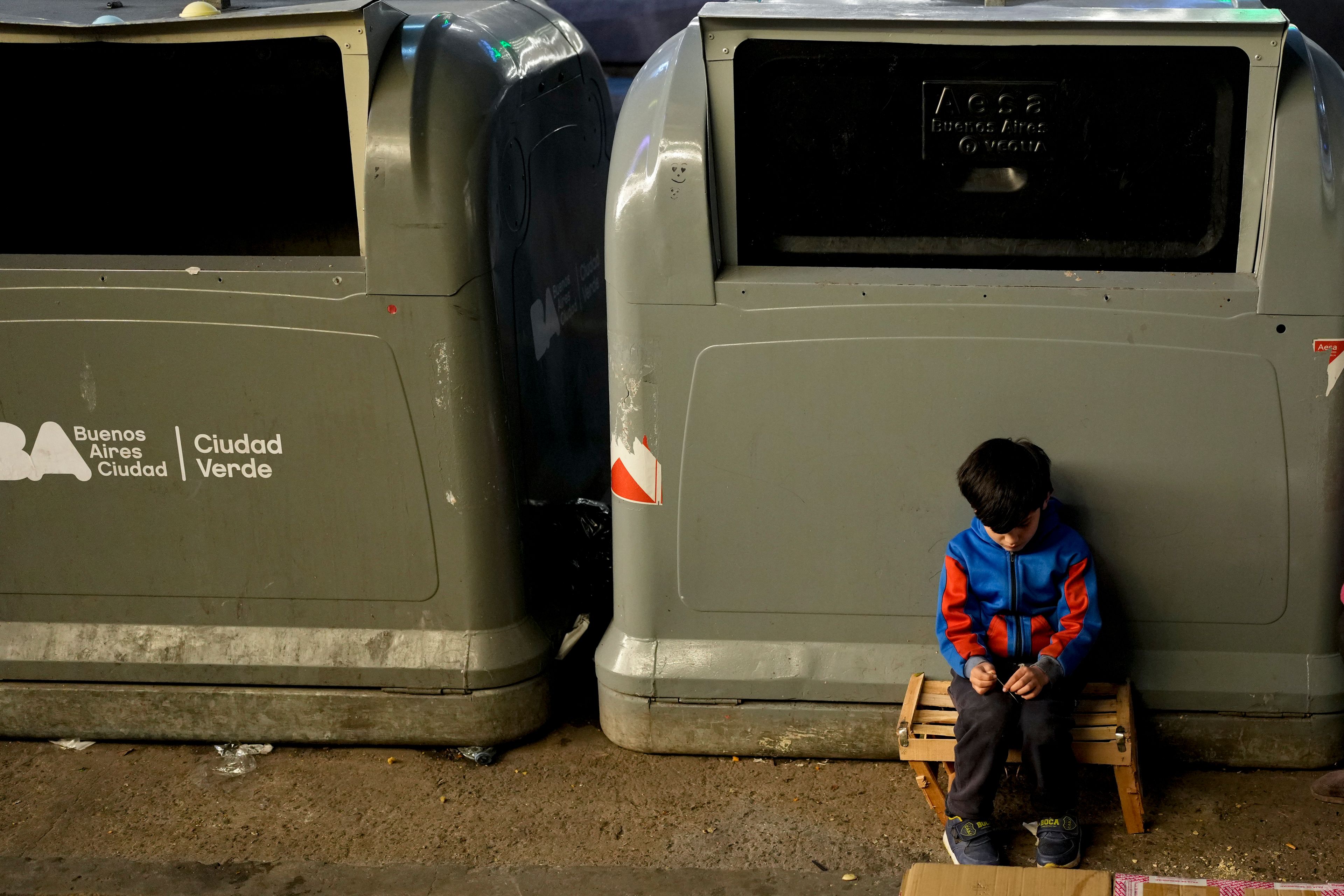 Tobias sits on the sidewalk next to public trash containers as his mother Elena Gonzeva sells handkerchiefs in the streets of Buenos Aires, Argentina, Tuesday, Sept. 3, 2024. (AP Photo/Natacha Pisarenko)