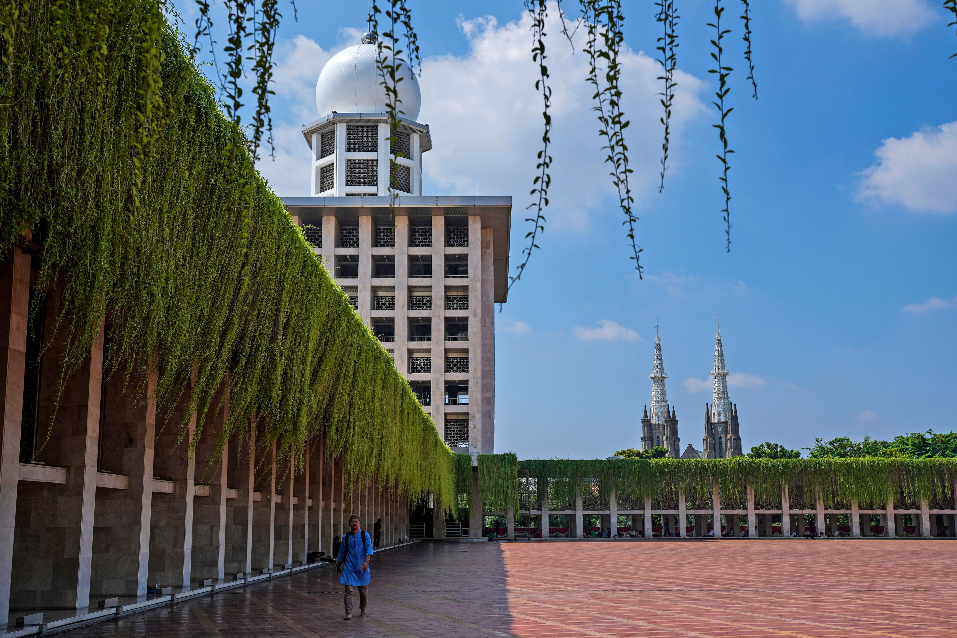 A man walks in the shade at Istiqlal Mosque as the spires of the Our Lady of The Assumption Cathedral are seen in the background, in Jakarta, Indonesia, Friday, Aug. 9, 2024.