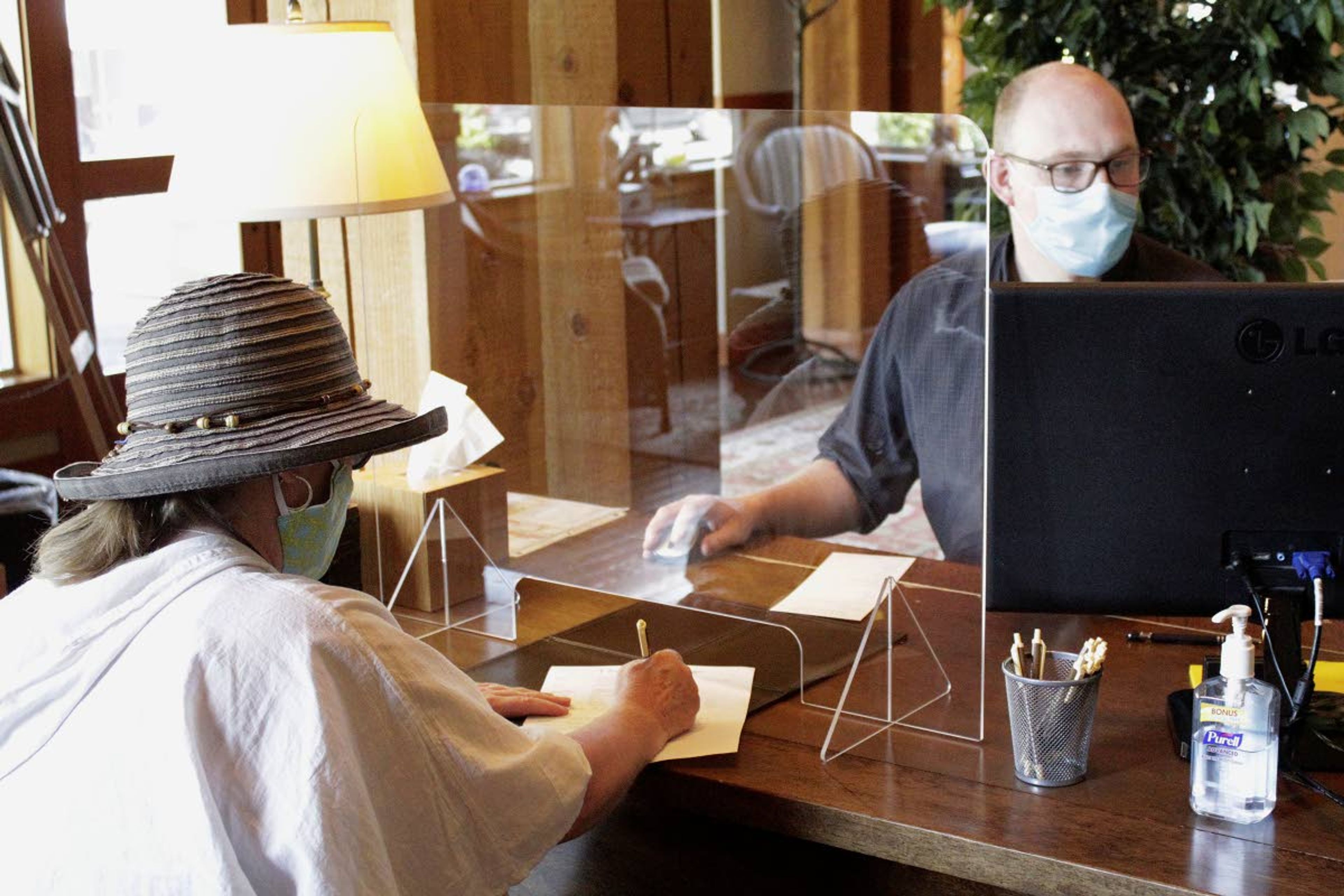 In this May 28 photo, hotel employee Daniel Mathre, right, helps a guest from behind protective plastic at The Ocean Lodge in Cannon Beach, Ore. The Ocean Lodge welcomed guests back starting last week after an extended shut-down of all hotels and vacation rentals in the small beach town to prevent the spread of the new coronavirus.