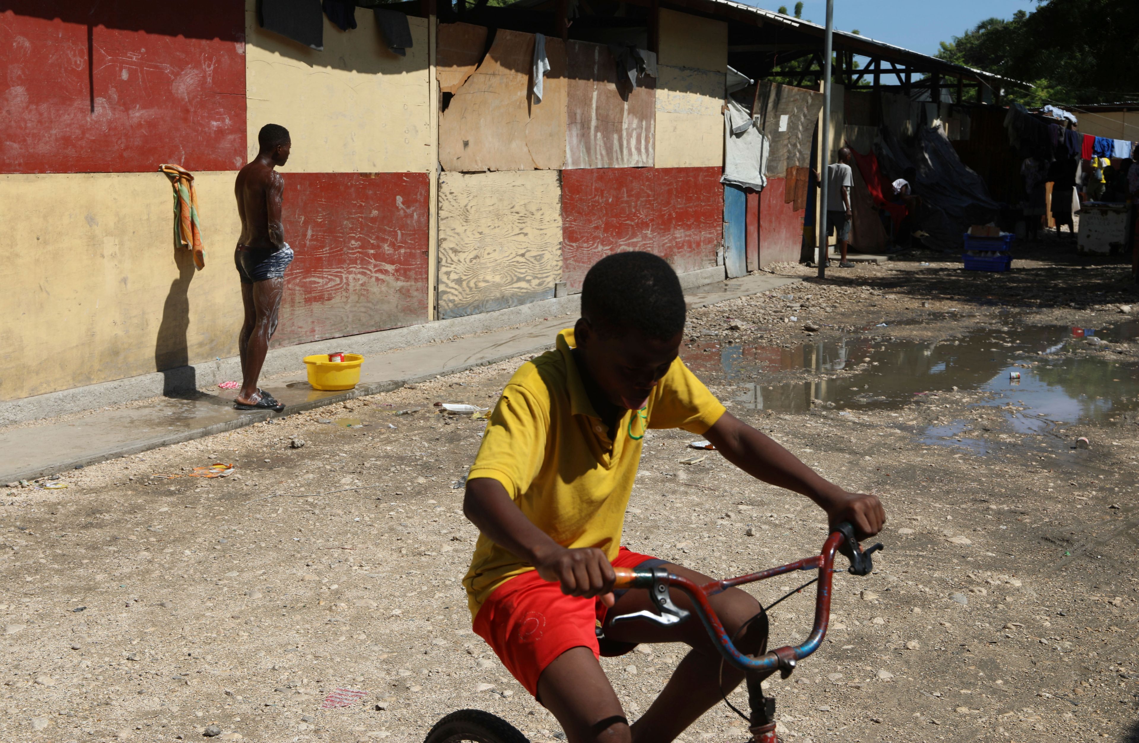 A youth cycles past a man taking a bucket bath at a school where people displaced by gang violence have been living for over a year in Port-au-Prince, Haiti, Sept. 20, 2024. (AP Photo/Odelyn Joseph)