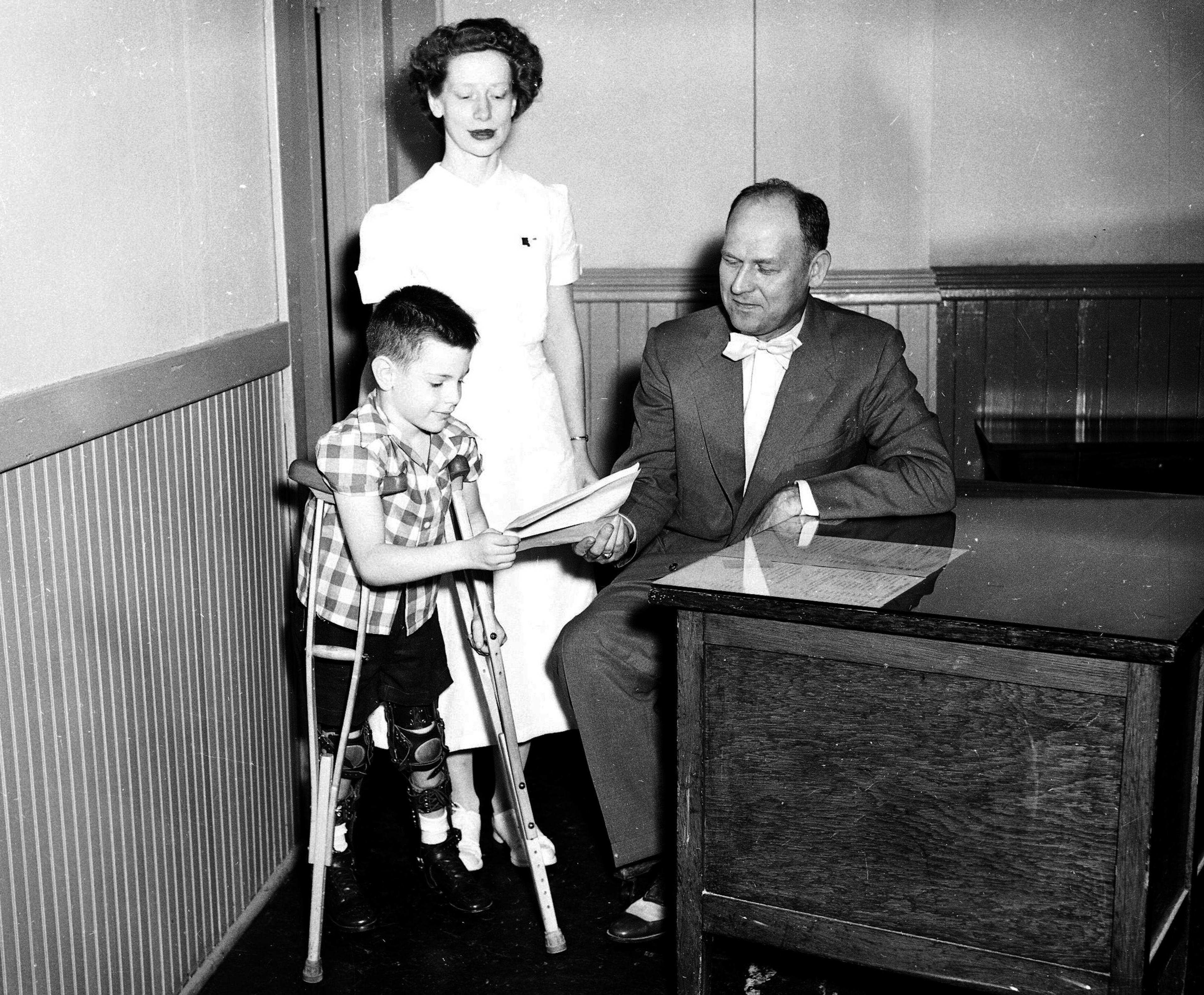 David Faris, 6, son of Mr. and Mrs. Fred Faris of Cape Girardeau, who was stricken with polio in August 1952, receives a copy of a proclamation from Mayor Narvol Randol setting aside the two weeks Aug. 6 to 30, 1954, as the emergency March of Dimes period in Cape Girardeau. The youngster is accompanied by Louise Steimle of the physical therapy department of the polio center, Saint. Francis Hospital.