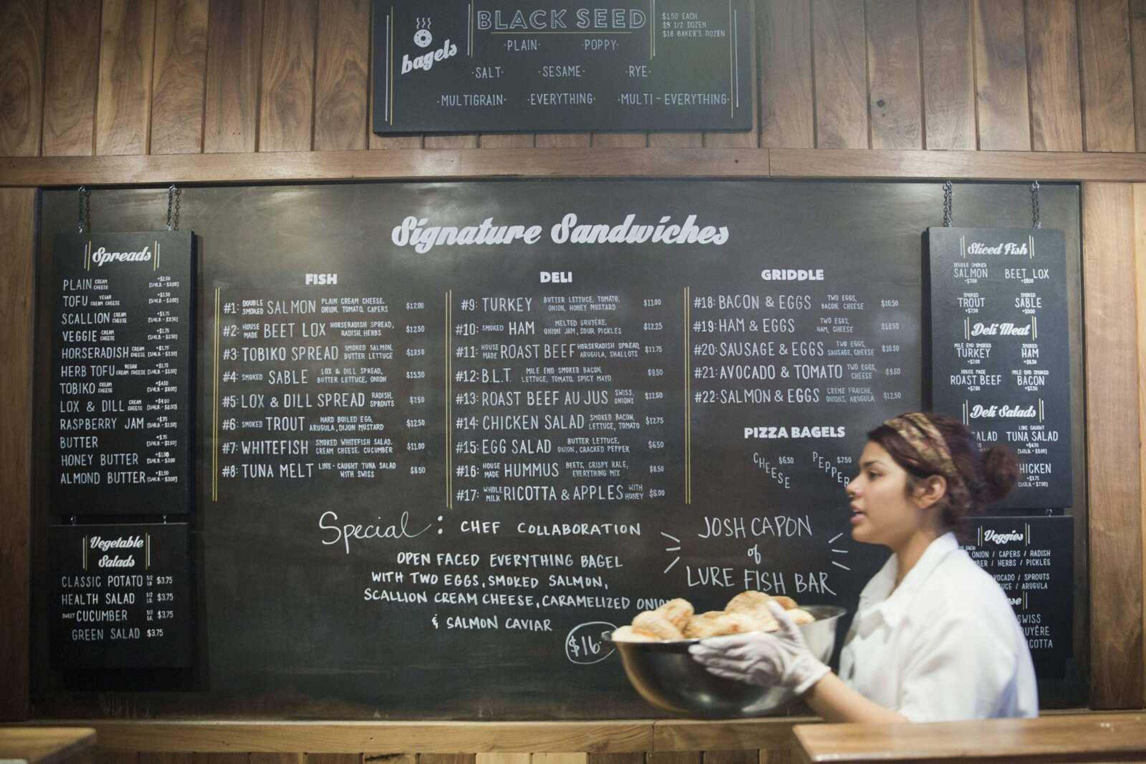 An employee of the Black Seed bagel shop walks freshly baked bagels past the store's menu in the East Village neighborhood of New York on Friday, May 27. (AP Photo/Mary Altaffer)