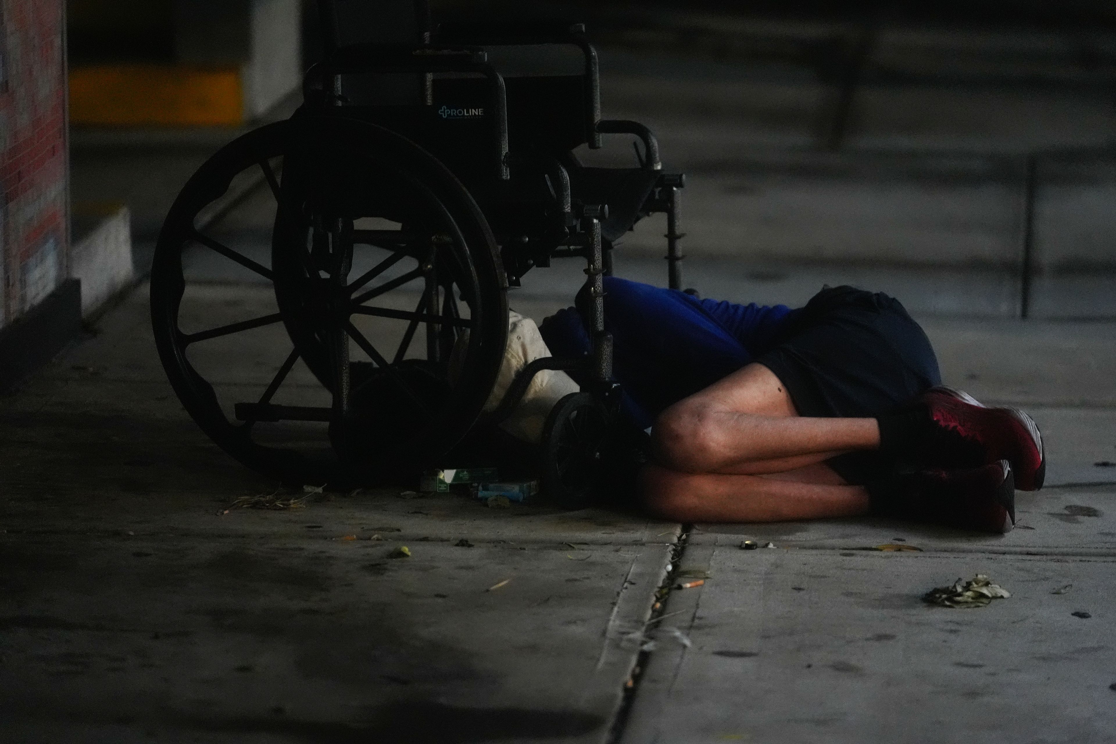 A homeless person sleeps under a wheelchair alongside a parking garage in deserted downtown Tampa, Fla., during the approach of Hurricane Milton, Wednesday, Oct. 9, 2024. (AP Photo/Rebecca Blackwell)