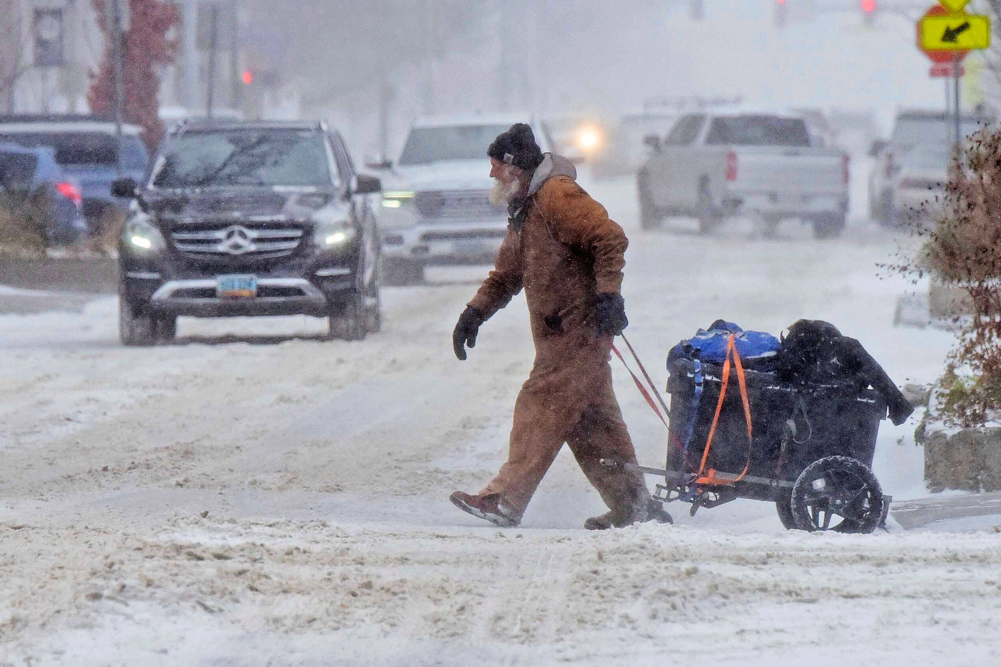 Chuck Benjamin pulls a cart with his dog named Shadow across Broadway Avenue as snow falls in downtown Bismarck, N.D., Wednesday morning, Nov. 20, 2024, in Bismarck, N.D. (Tom Stromme/The Bismarck Tribune via AP)