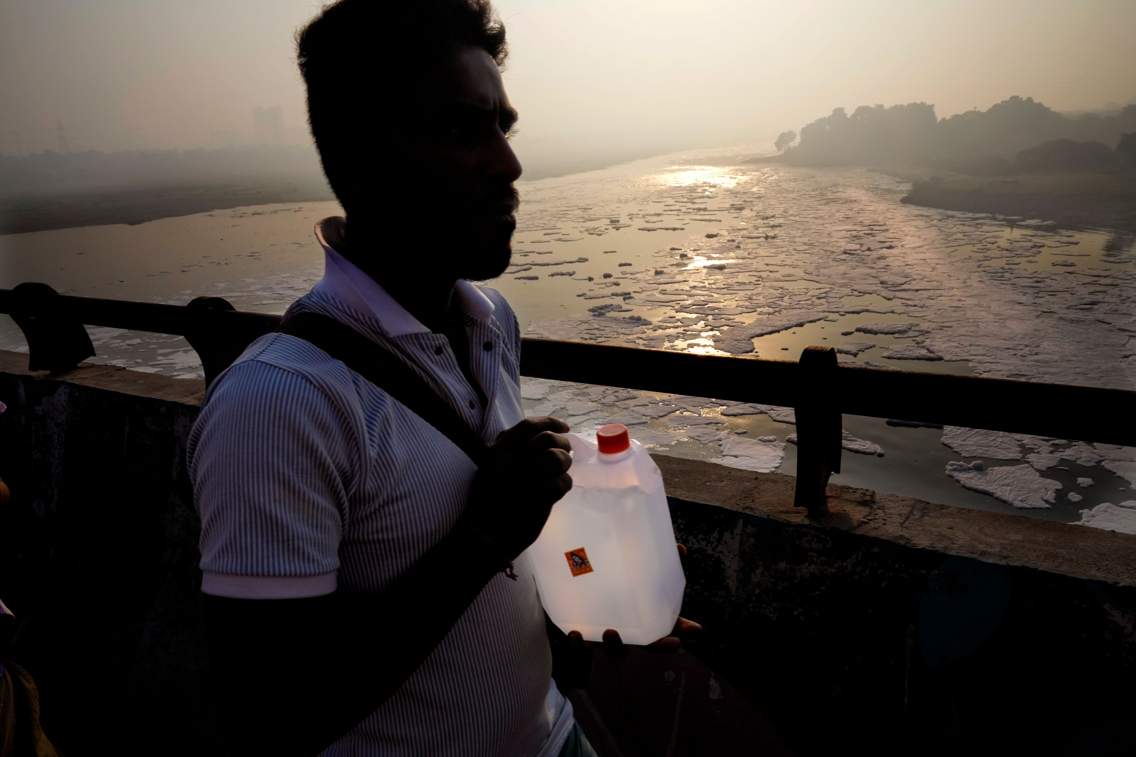 A person carries sacred Ganges river water in a container as he walks across the bridge over the river Yamuna filled with toxic foams in New Delhi, India, Tuesday, Oct. 29, 2024. (AP Photo/Manish Swarup)