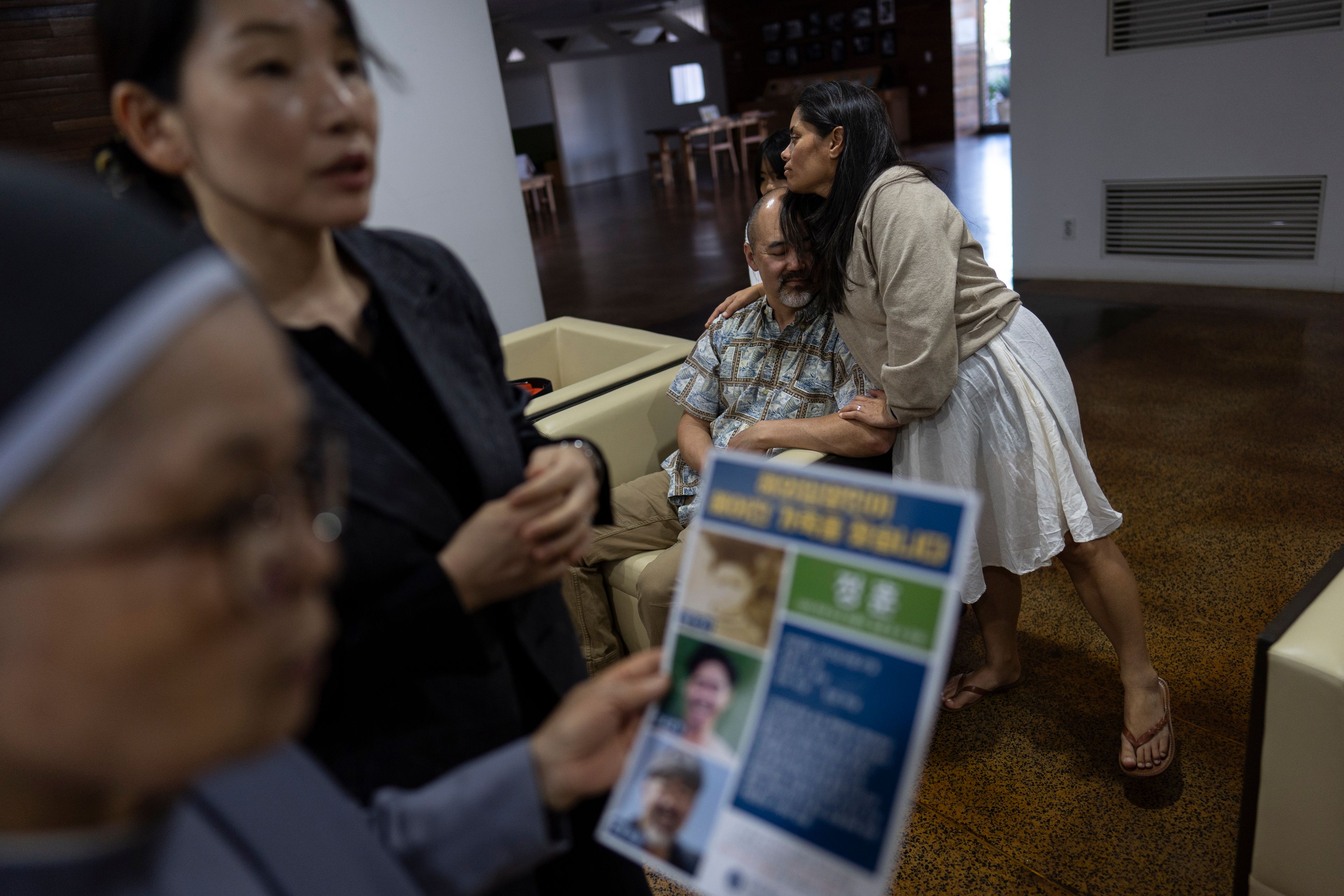 Kenneth Barthel, who was adopted by a single parent in Hawaii at 6 years old, is hugged by his wife, Napela, at the Sisters of Mary in Busan, South Korea, Friday, May 17, 2024. In the foreground, Sister Bulkeia, left, and Paek Kyeong-mi from Global Overseas Adoptees' Link discuss a flyer designed to uncover the details of Barthel's early life and find his birth family. (AP Photo/Jae C. Hong)