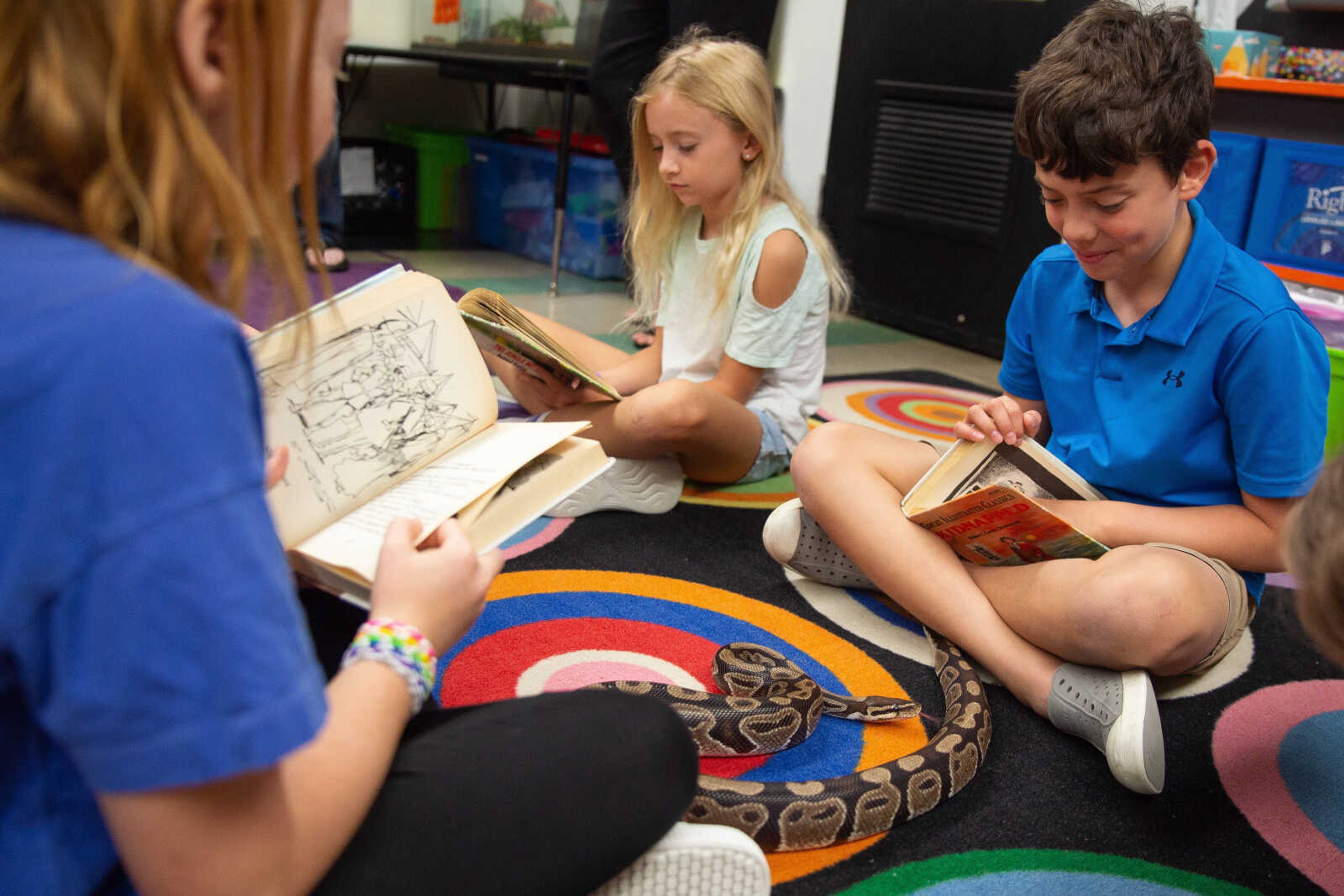 John Stewart Humphrey smiles as Monty the snake slithers around his feet as he sits reading with fellow classmates, Annabel Waggoner and Charis Cheek.