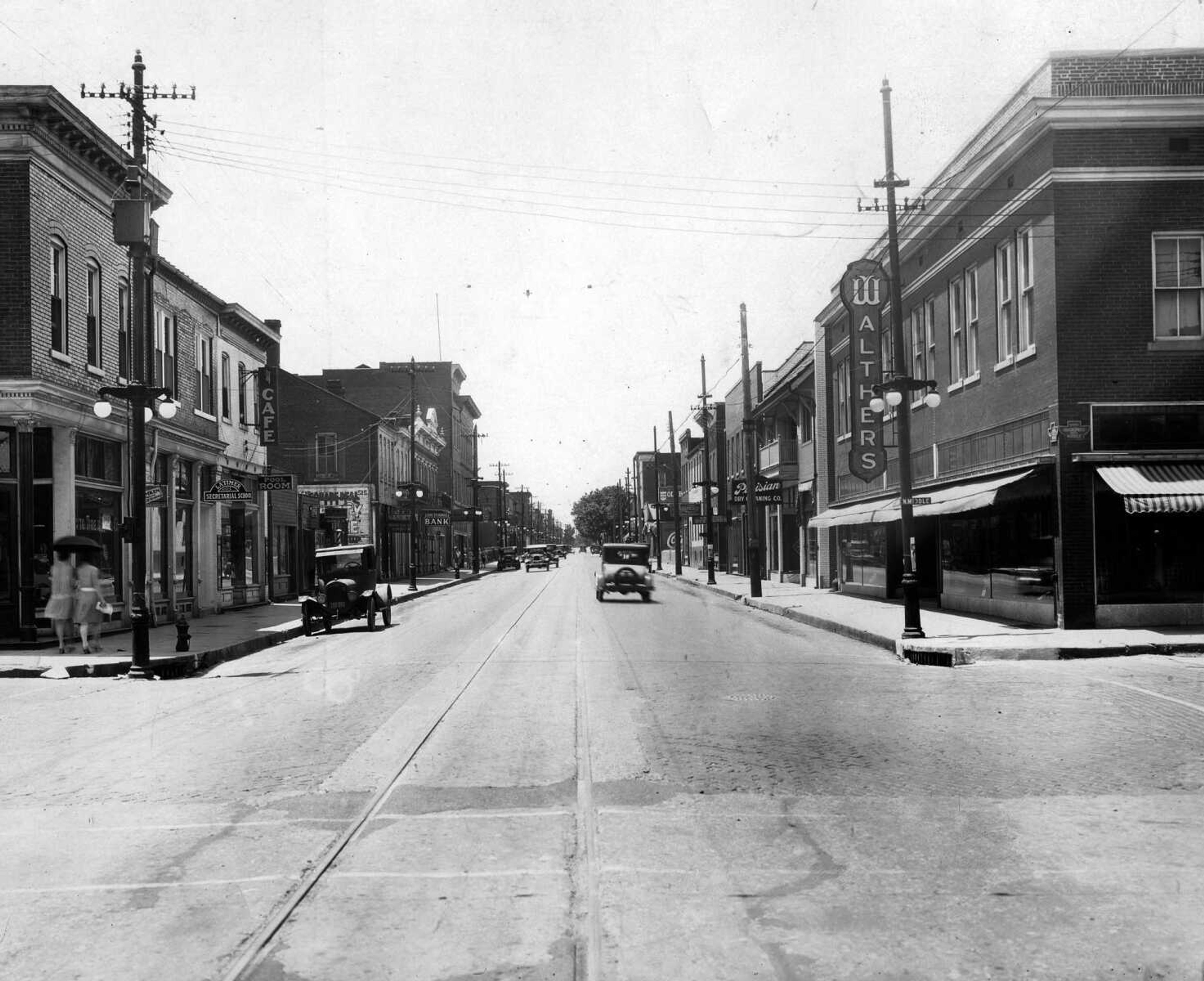 An umbrella shades two women walking west along Broadway in this undated picture, taken at the Middle Street intersection. Some of the businesses on the south side of the block are Auto Tire and Parts, Latimer Secretarial School, Square Deal Variety Store, Cape Exchange Bank and Bergmann Grocery. On the other side, some of the shops are Walther's Furniture Store, Parisian Dry Cleaning and Western Dye Works.