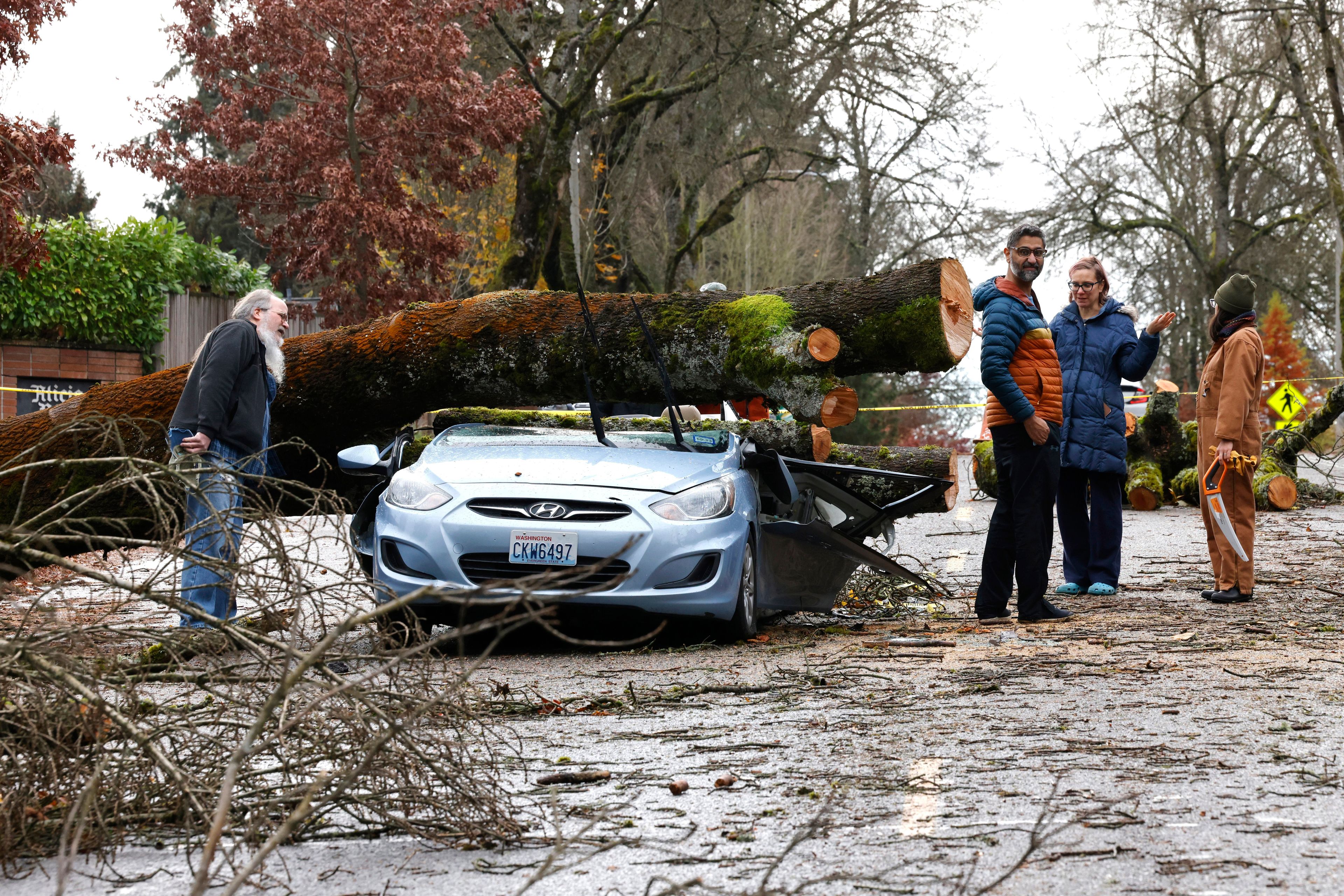 Neighbors look at the remains of small car that had a tree fall on it in the aftermath of a "bomb cyclone" on 35th Avenue Northeast after severe weather hit last night, in Seattle, Wednesday, Nov. 20, 2024. (Karen Ducey/The Seattle Times via AP)