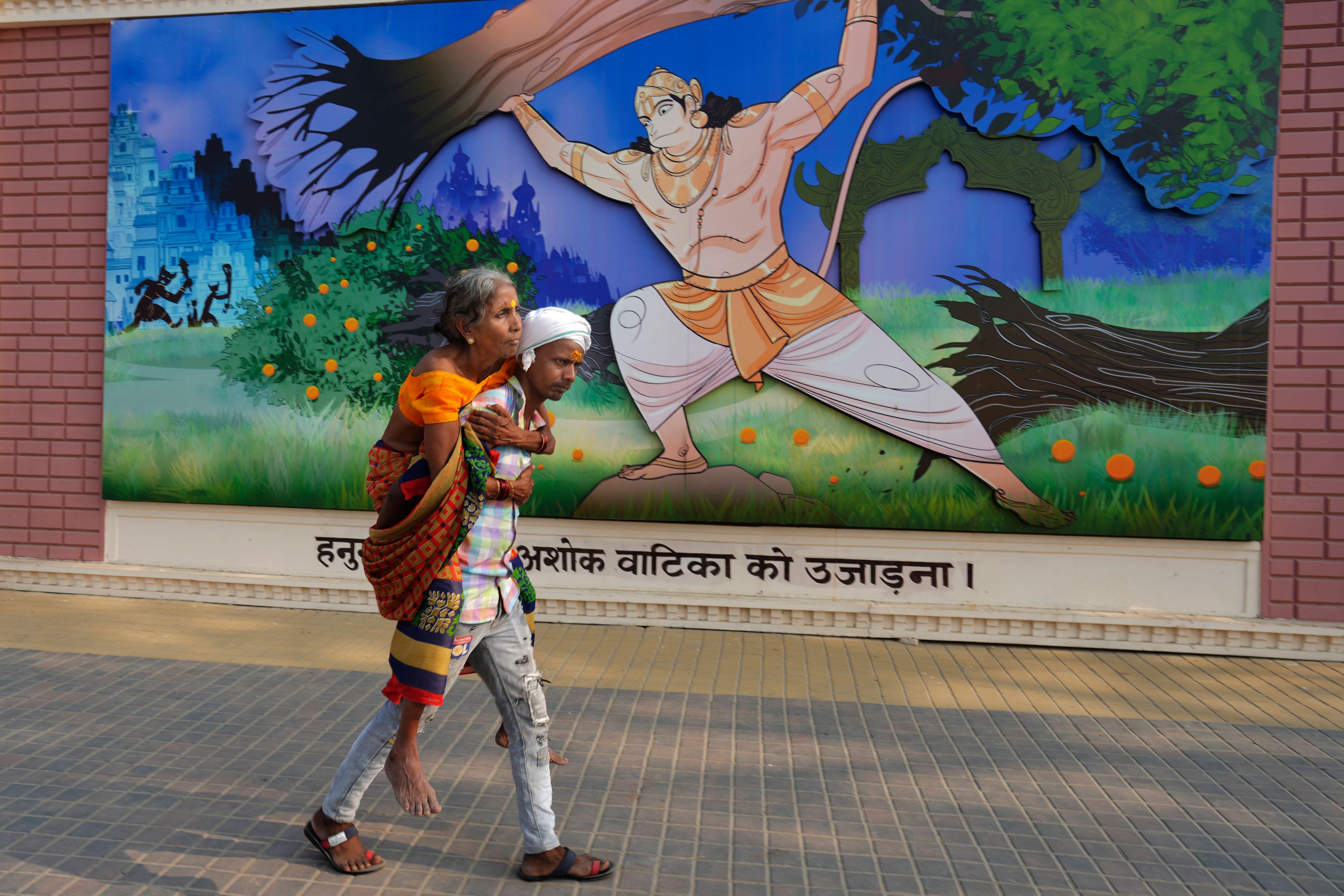 A man carries his elderly mother as he walks past a wall mural showing Hanuman, a Hindu deity, on the morning of Deepotsav celebrations, an event organized by the Uttar Pradesh state government on the eve of Diwali, in Ayodhya, India, Wednesday, Oct. 30, 2024. (AP Photo/Rajesh Kumar Singh)