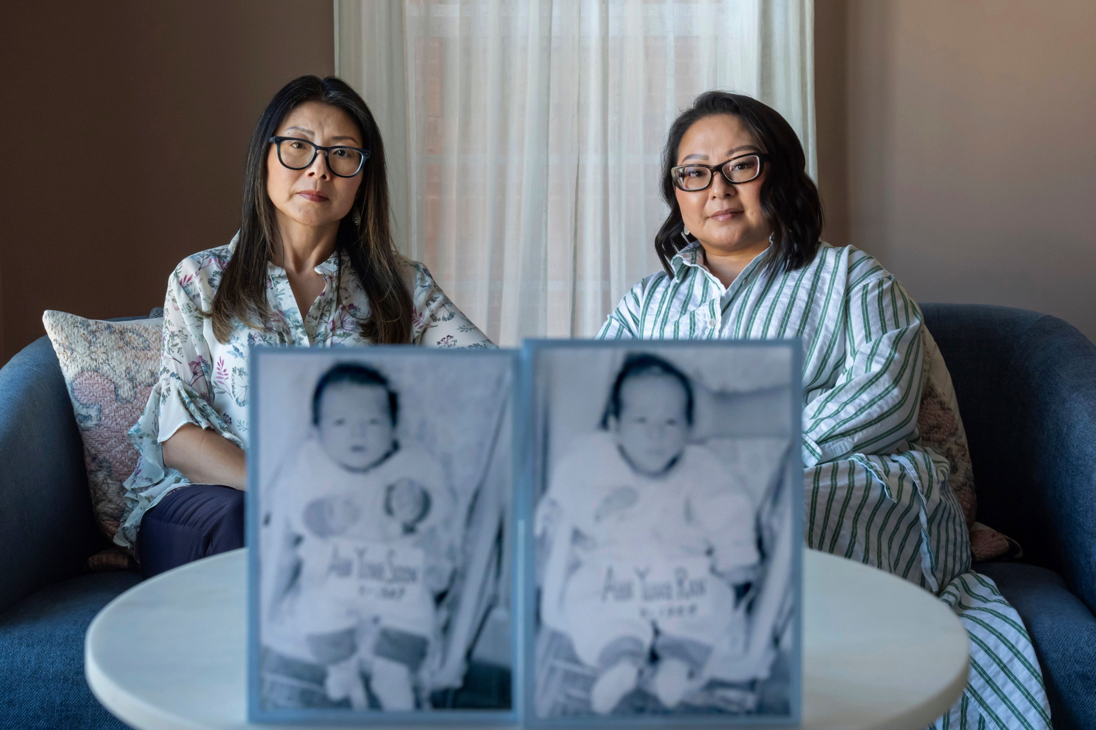 Dee Iraca, left, and her twin sister, Becca Webster, sit behind their baby photos from before they were adopted out of South Korea to a family in the United States, Saturday, April 6, 2024, at Iraca's home in Davidson, N.C. After all, they ended up happy in America. Yet their happiness was built on an injustice that hurt thousands, including their birth father and were too late to meet their mother. (AP Photo/David Goldman)