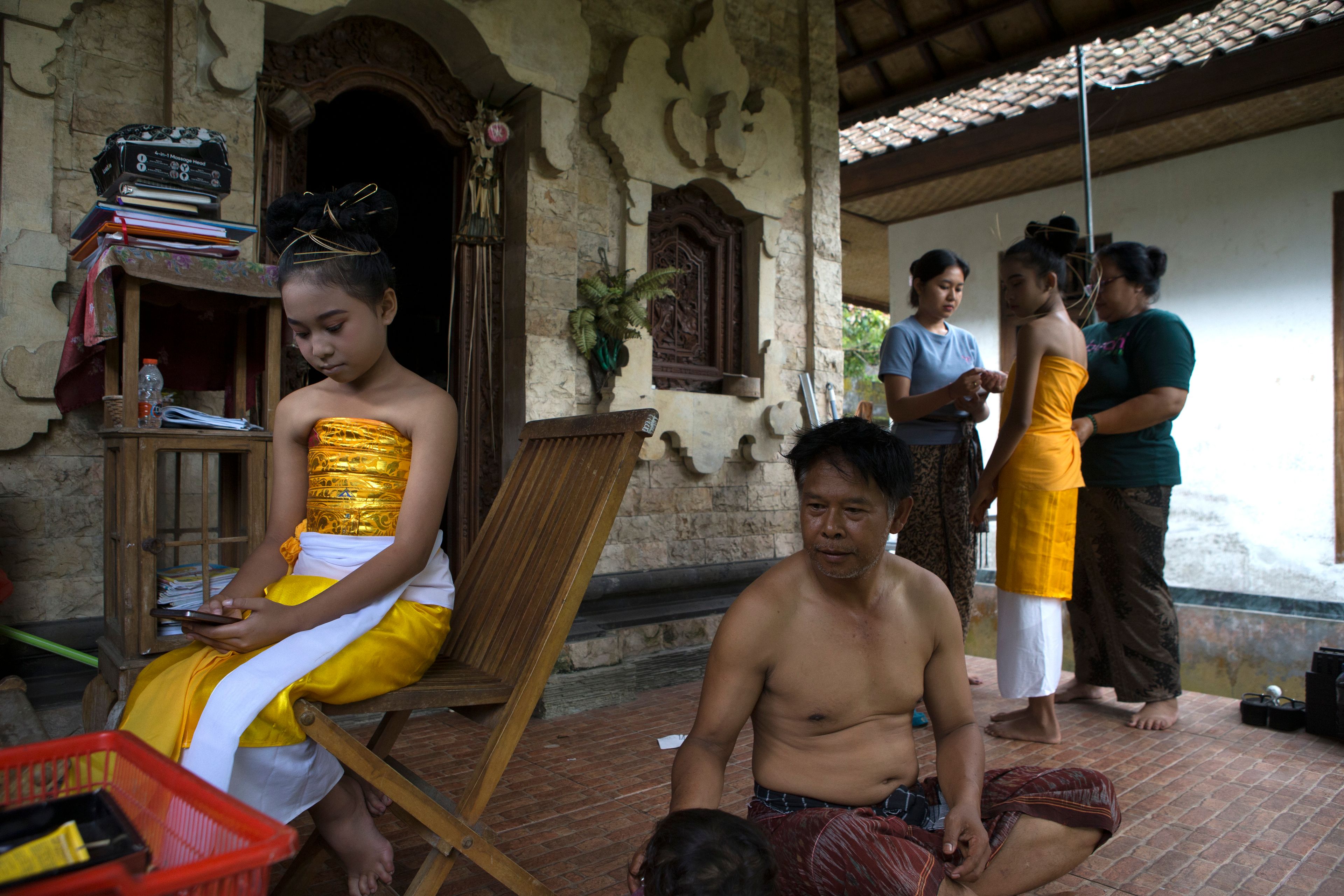 Ketut Nita Wahyuni, 11, looks at her phone with her father Nyoman Subrata sitting beside her, as mother Kadek Krisni, right, and sister Intan Wahyuni, in blue, dresses up her cousin Rina Lestianti before the Rejang Pucuk Hindu rituals at Geriana Kauh village, Karangasem, Bali, Indonesia on Thursday, Nov. 21, 2024. (AP Photo/Firdia Lisnawati)