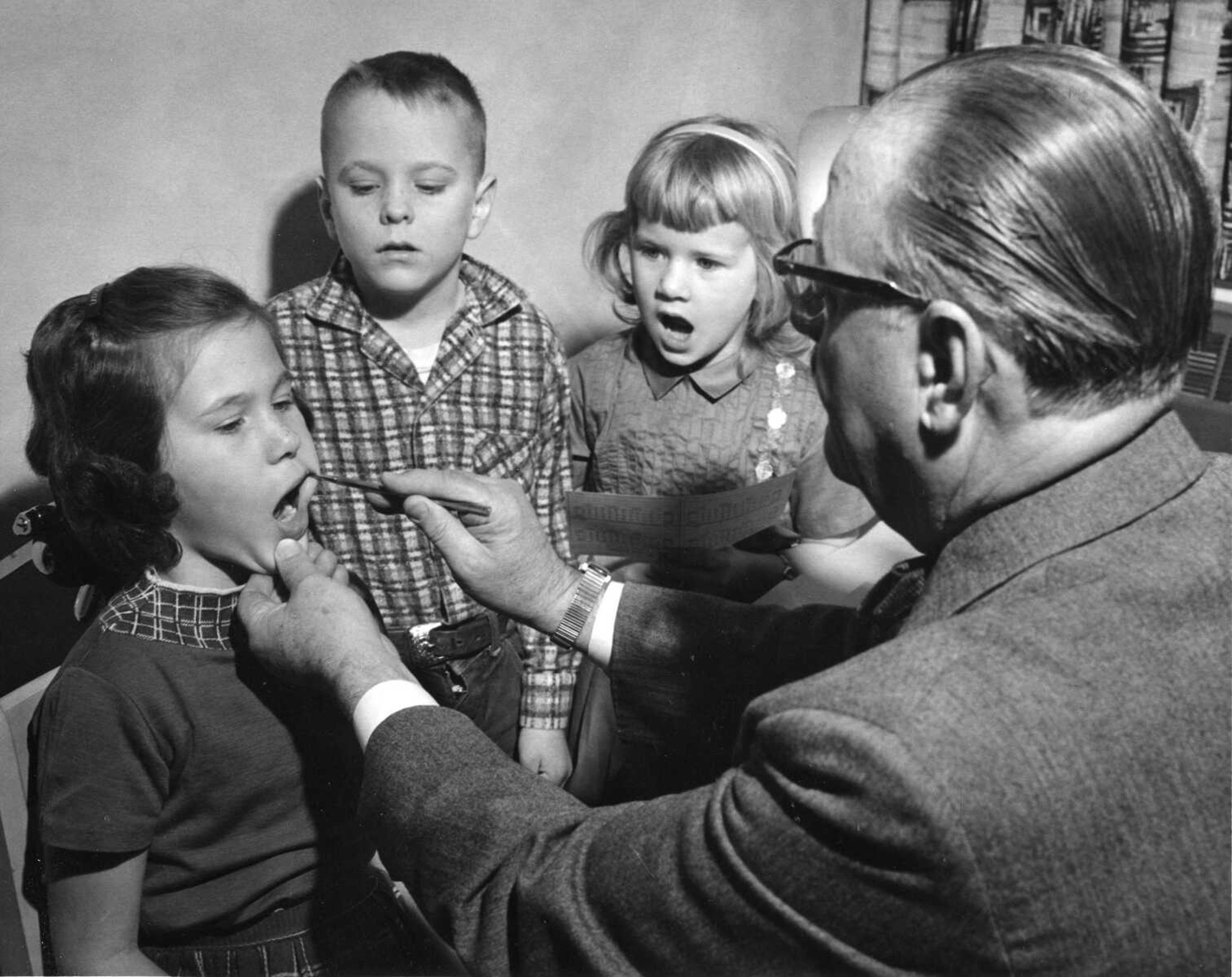 Published Thursday, Feb. 28, 1962
Joan Lynn Baker, daughter of Mrs. Mary Lou Baker, 1241 Marilyn Drive, received a sympathetic reaction from Vickie Sue Bailey, right, daughter of Mr. and Mrs. Floyd C. Bailey, 1300 Victoria, at the dental check at Alma Shrader School this morning. Dr. Richard Brown, president of the District Dental Study Club,, is making the examination and Steven Paul Berkbigler, son of Mr. and Mrs. Ladelle Berkbigler, 1712 Westwood, is giving a critical glance as he awaits his turn. The pupils are all 6 years old and in the first grade. The examinations were carried out in all elementary schools and coincided with National children's Dental Health Week. (Missourian photo by G. D. Fronabarger)