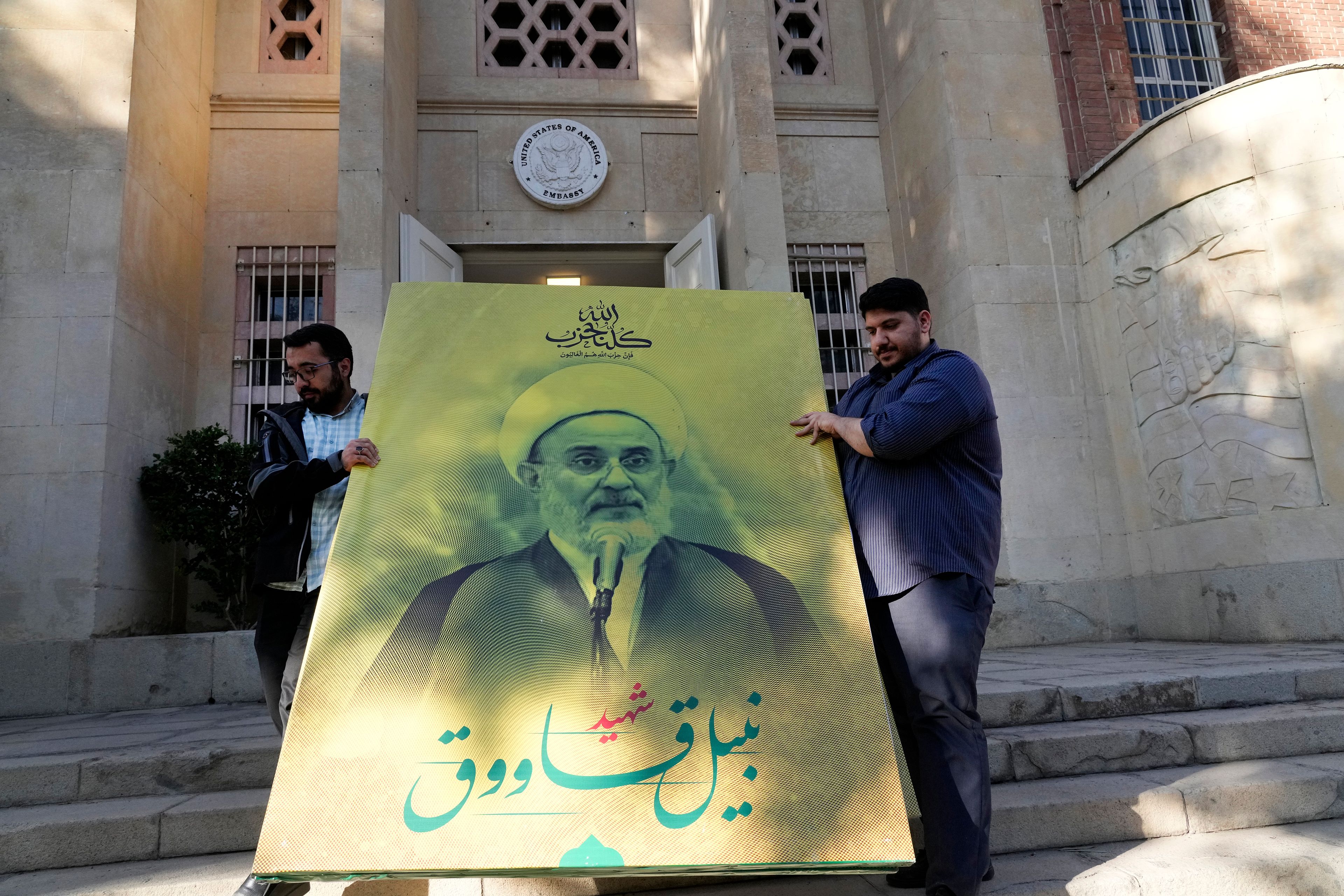 Two men carry a banner of slain deputy head of Hezbollah's Central Council Nabil Kaouk, who was killed in an airstrike by Israel in late September, at the entrance of the former U.S. Embassy, which has been turned into an anti-American museum, in Tehran, Iran, Tuesday, Oct. 22, 2024. (AP Photo/Vahid Salemi)