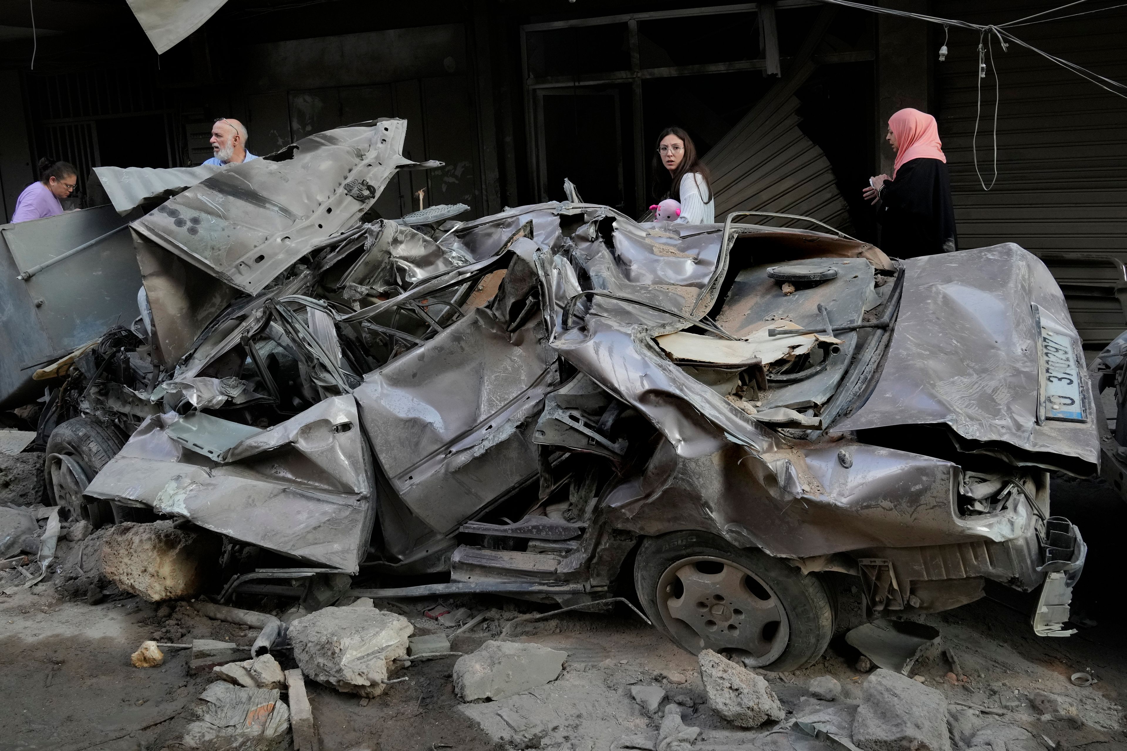 People pass by a destroyed car at the site of Thursday's Israeli airstrike, in Beirut, Lebanon, Friday, Oct. 11, 2024. (AP Photo/Hussein Malla)