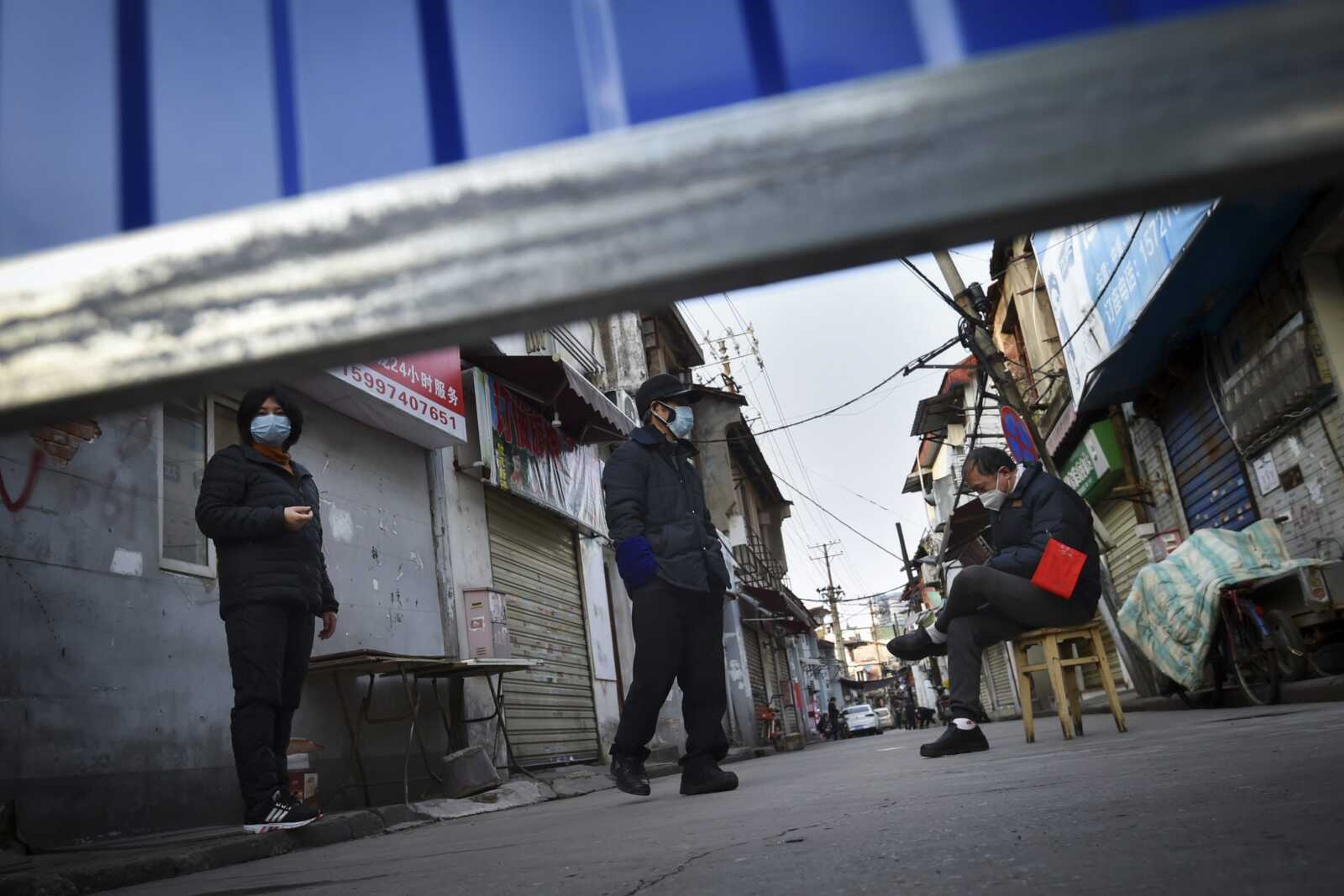 A volunteer sits on a chair keeping guard as residents stand near the barricades blocking a residential area Sunday in Wuhan in central China's Hubei province. Warning that China's virus epidemic is "still grim and complex," President Xi Jinping called Sunday for more efforts to stop the outbreak, revive industry and prevent the disease from disrupting spring planting of crops.