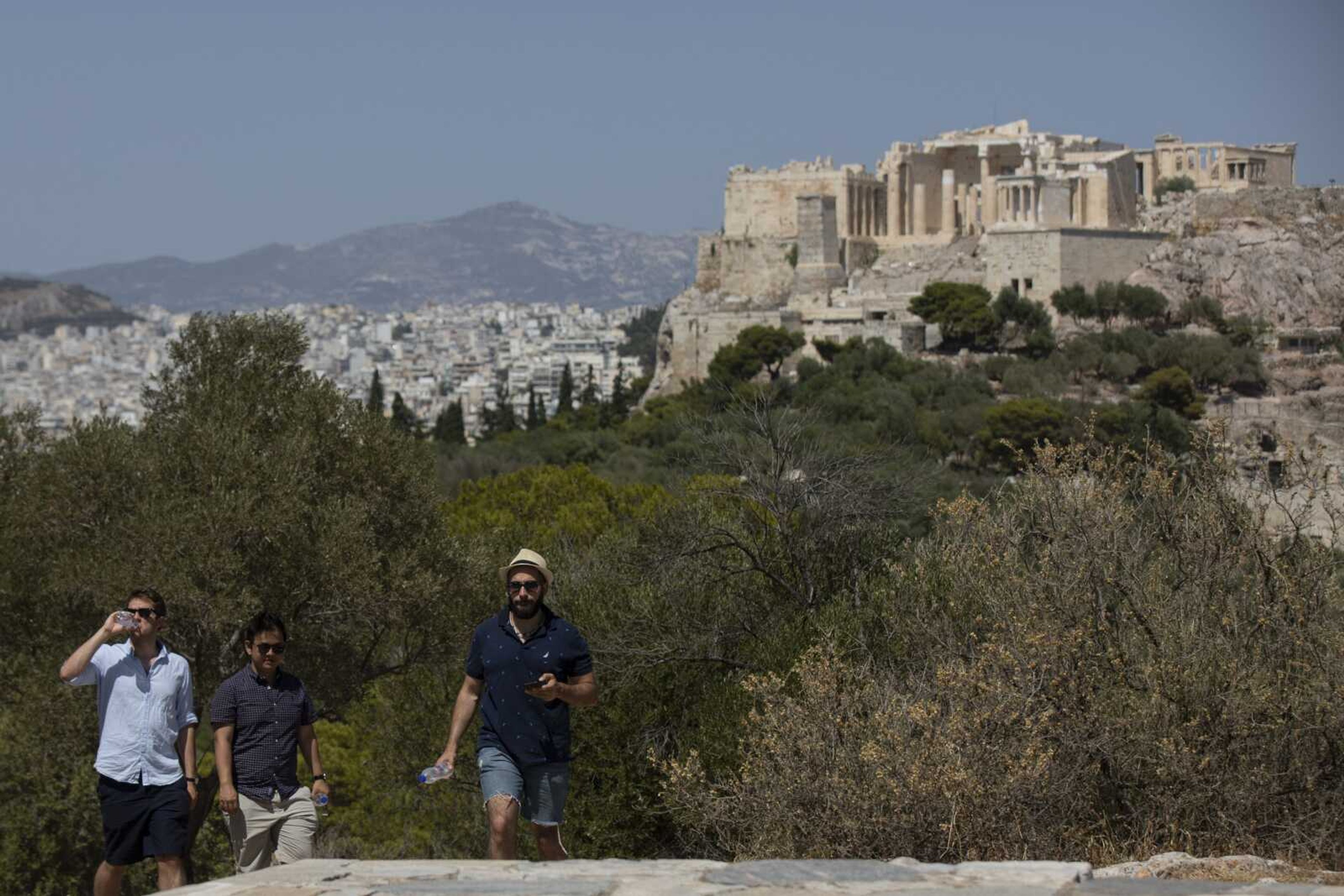 A tourist drinks water while walking with others at Filopappos hill as at the background is seen the ancient Acropolis hill during a hot day Thursday in Athens. Greece's most famous archaeological site, the Acropolis in Athens, has shut down to visitors for four hours because of hot weather in the capital.