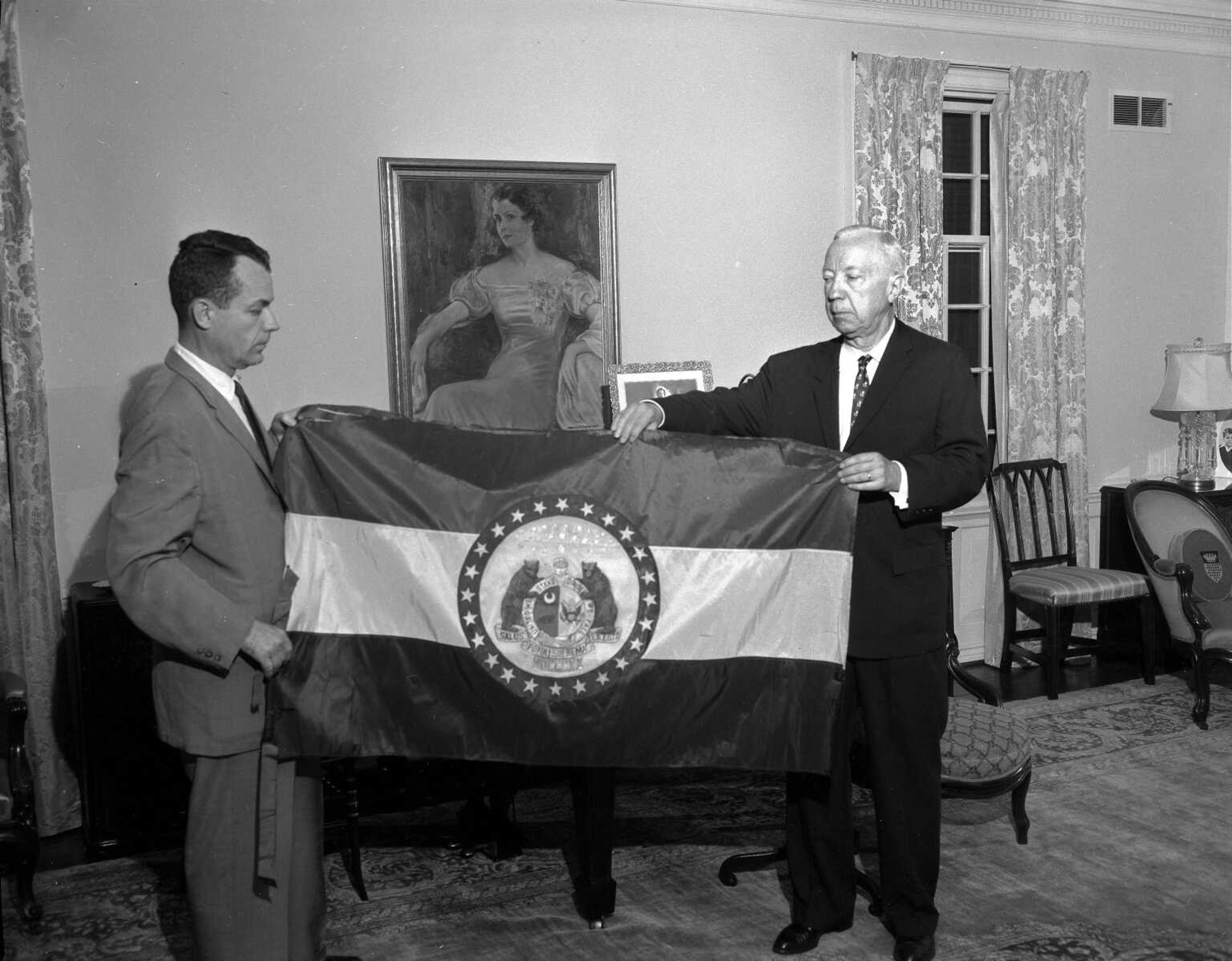 Published Wednesday, June 14, 1961
Cape Girardeau today lost the distinction of being home for the original Flag of Missouri. Today it was presented in formal ceremonies at a joint session of the Senate and House in Jefferson City to the State of Missouri to become one of its cherished historic items. Shown in a picture before the flag was taken to Jefferson City Tuesday are a grandson, Jack Oliver, and a son, Allen L. Oliver, of Mrs. R.B. Oliver, who designed the flag and of whom an early day portrait appears in the background.
(Photo by G.D. "Frony" Fronabarger, Cape Girardeau Southeast Missourian)