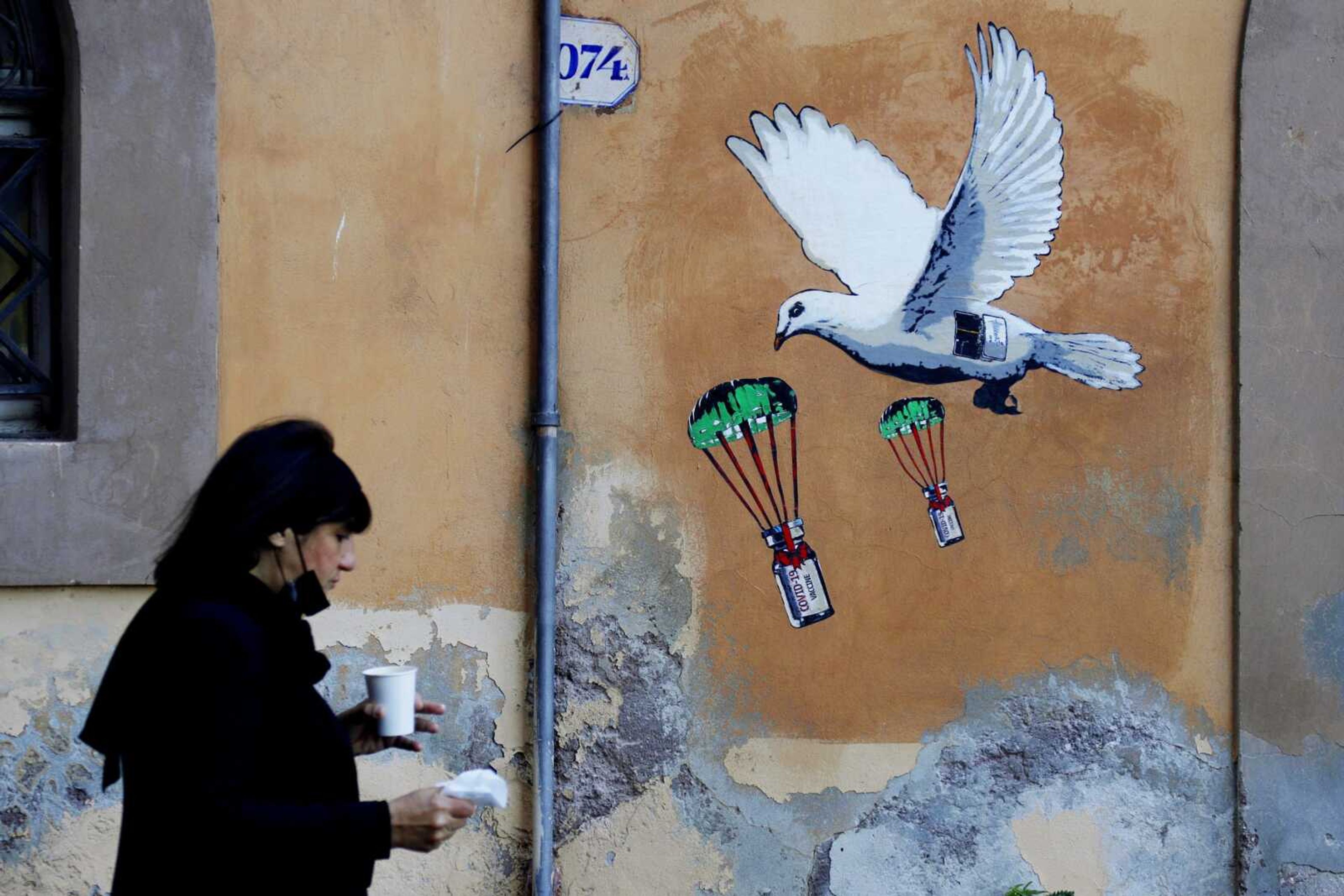 A woman walks past a mural depicting a white dove parachuting COVID-19 vaccine vials, posted near the Italian Health Ministry Headquarters, on Sunday in Rome.