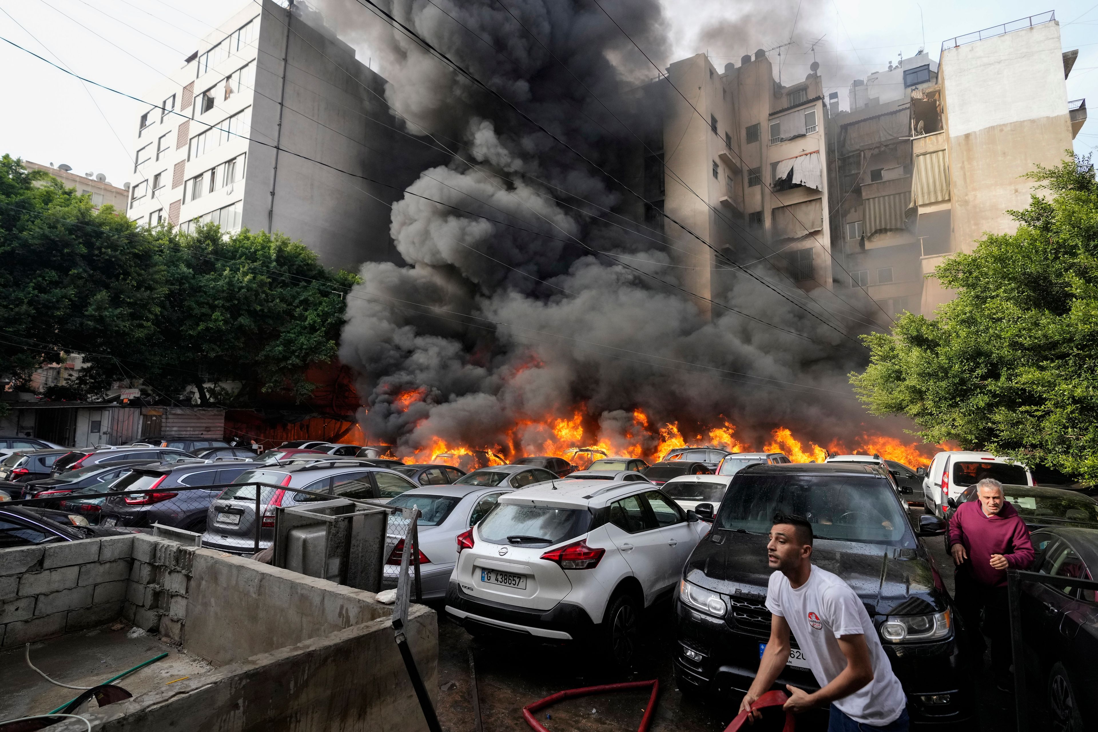 A man runs as a civil defense worker tries to extinguish a fire caused by a giant diesel tank which ignited, burning more than a dozen cars in a parking lot and damaging a building nearby, in a residential Beirut neighborhood, Lebanon, Saturday, Nov. 9, 2024. (AP Photo/Hassan Ammar)