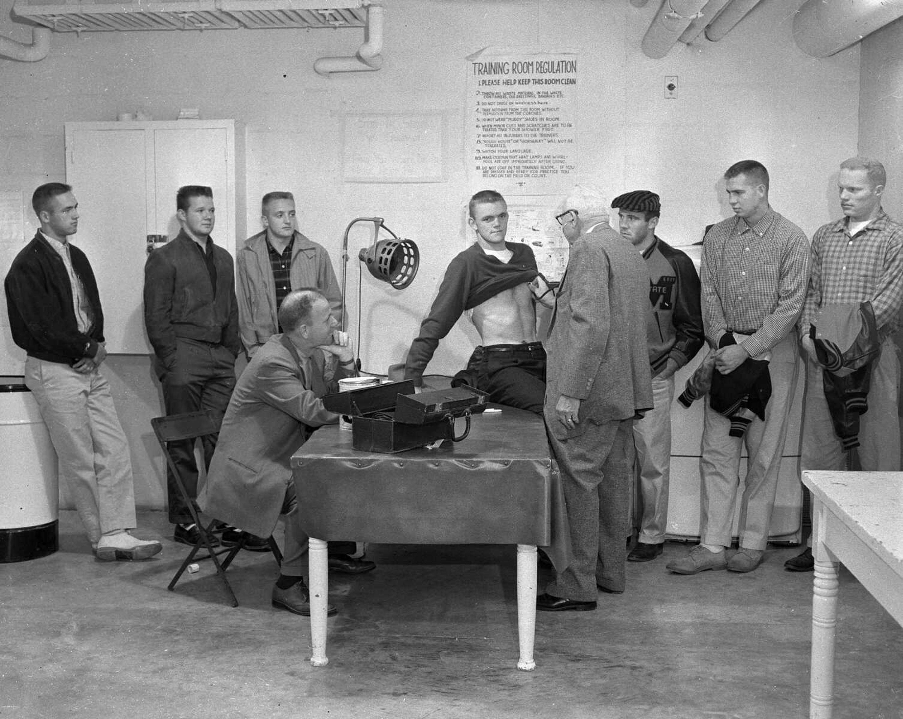 Oct. 25, 1957 Southeast Missourian.
Little flu bug downs tough State College gridders. With 20 of the regular squad of 33 players ill in varying degrees from influenza, the State College Indians were forced to postpone their scheduled game Saturday at Rolla with the Missouri Miners, and this photo shows ailing gridmen receiving examination and treatment at the Houck Field House training room. From the left are Don Varner, Jerry McConnell, Jim Springs, Bill Stacy, Arlen Powell, Frank Czapla and John Nicholas. Seated is Coach Kenneth Knox, who is watching Dr. O.L. Seabaugh, college physician, examine Stacy. (G.D. Fronabarger/Southeast Missourian archive) [Bill Stacy would become the 12th president of Southeast Missouri State University from 1980-1989. Dr. Oda Lavinia Seabaugh practiced medicine in Cape Girardeau 43 years before ill health forced his retirement in 1959. He suffered a stroke in 1954 which resulted in limited practice until his retirement. He died at 85 on Dec. 9, 1970. Dr. Seabaugh was at the forefront when the Spanish influenza epidemic of 1918-1919 hit Cape Girardeau. He treated more than 100 members in the Student Army Training Corps at State College who were stricken, in addition to his private practice. All of his patients recovered.]