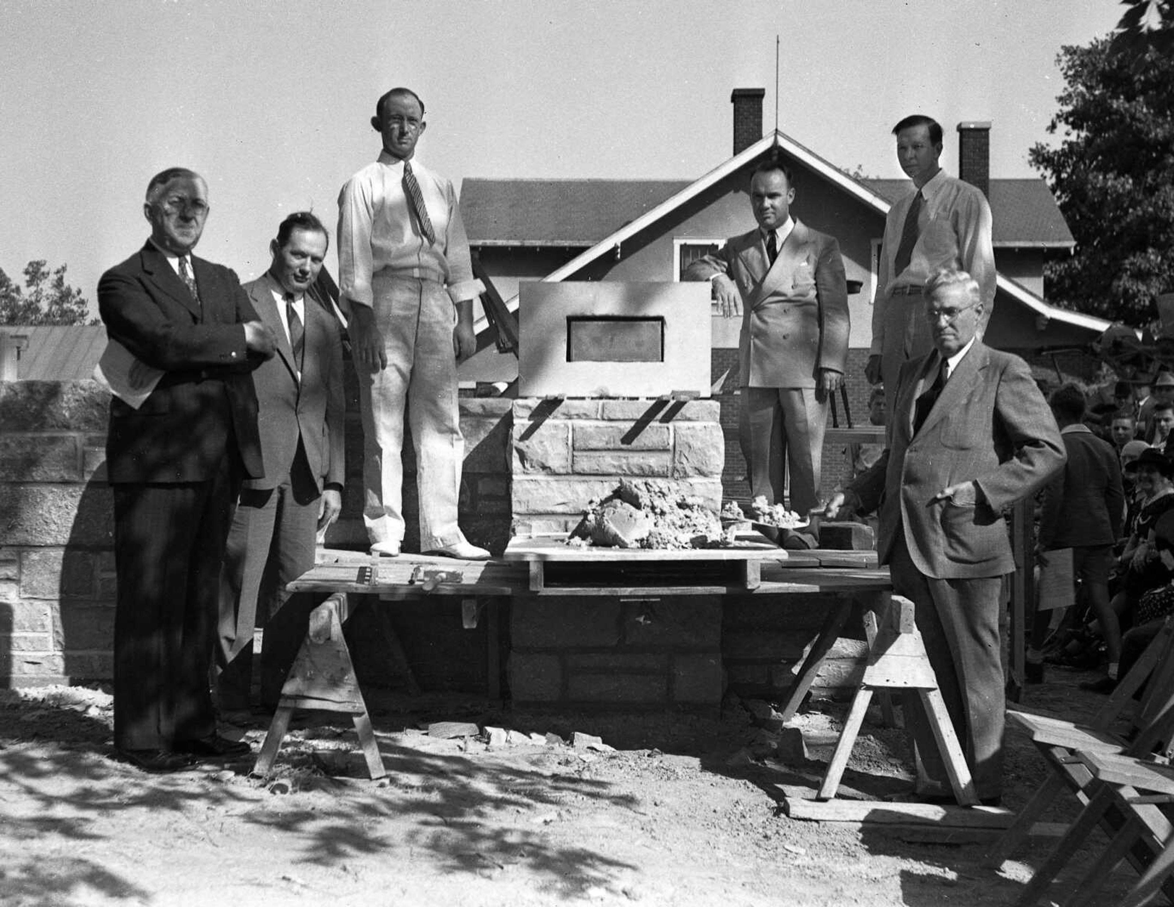 A group of serious-looking men are prepared to place a cornerstone for a stone building that's under construction. Do you recognize any of these men or the structure being built?