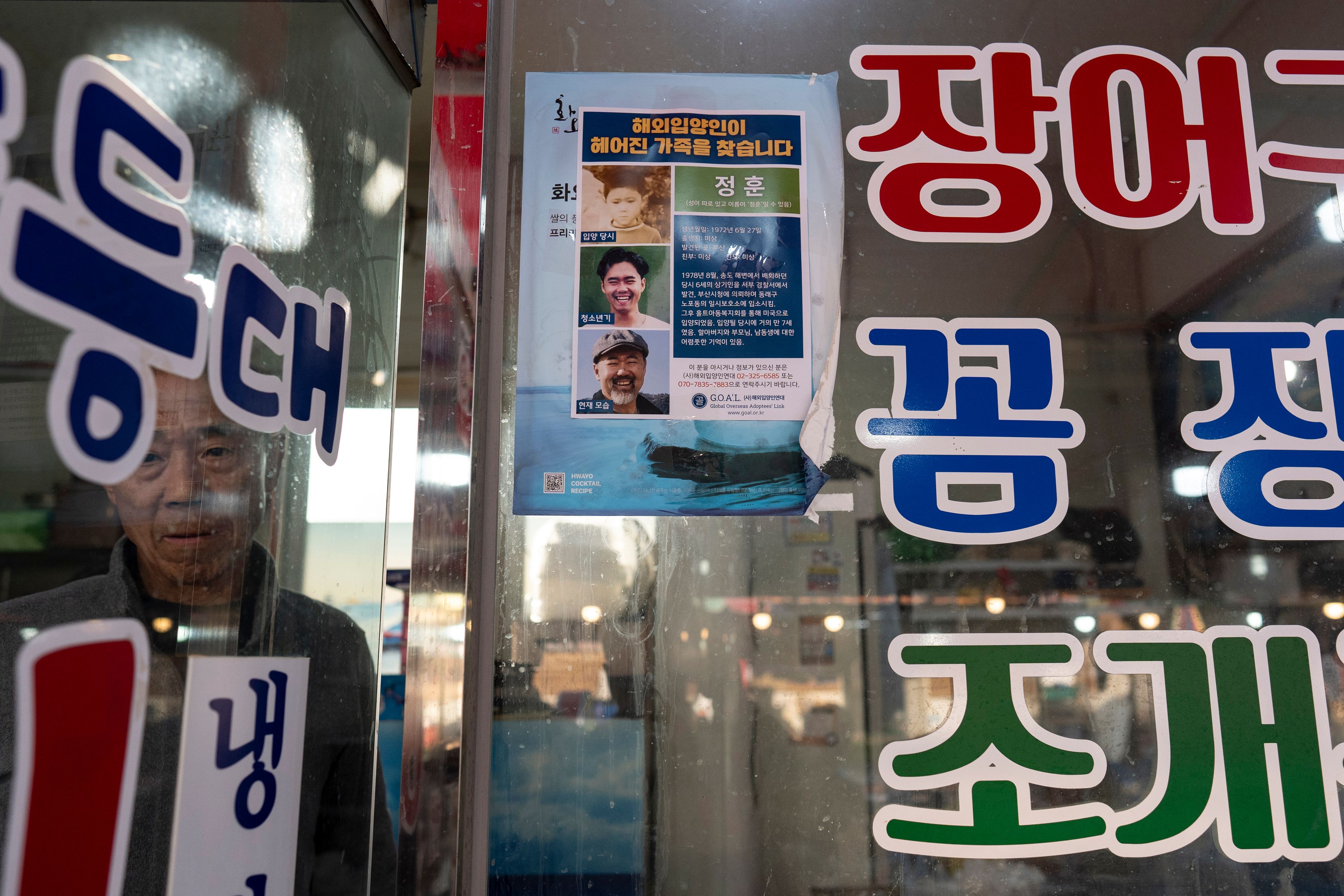 Restaurant owner Shin Byung-chul looks from behind a flyer he put up of Kenneth Barthel, who was abandoned in the area as a child and later adopted to Hawaii at 6 years old, at his restaurant in Busan, South Korea, Friday, May 17, 2024. Barthel has visited Korea four times to search for his birth family. (AP Photo/Jae C. Hong)