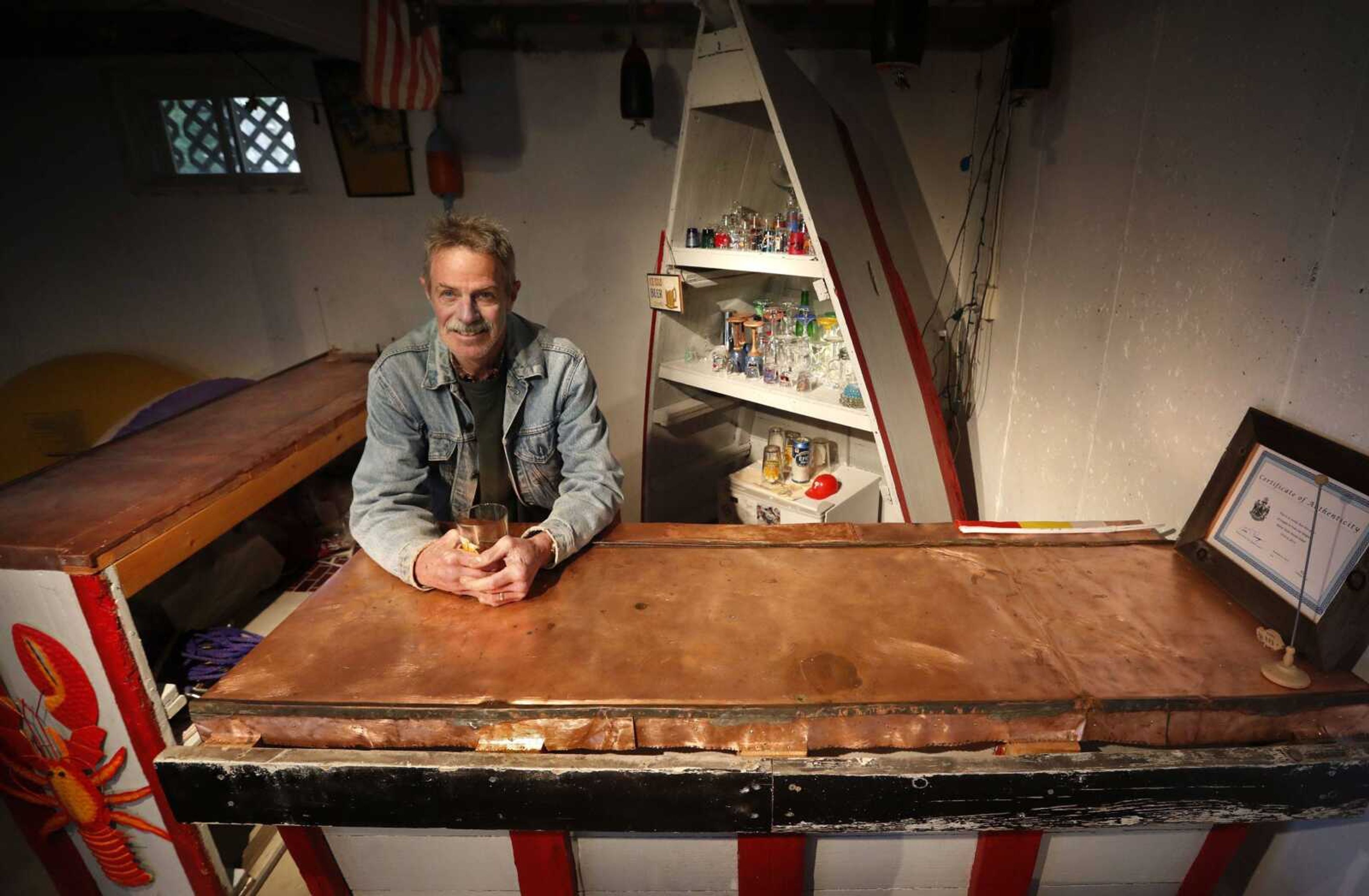 In this Nov. 2 photo, Glenn Adams poses behind a bar he made from parts of a sunken 18-foot boat and copper that once covered the dome at the Maine State House, at his camp in Richmond, Maine. The boat was cut into three pieces, two for the bars and one for shelving in the background.