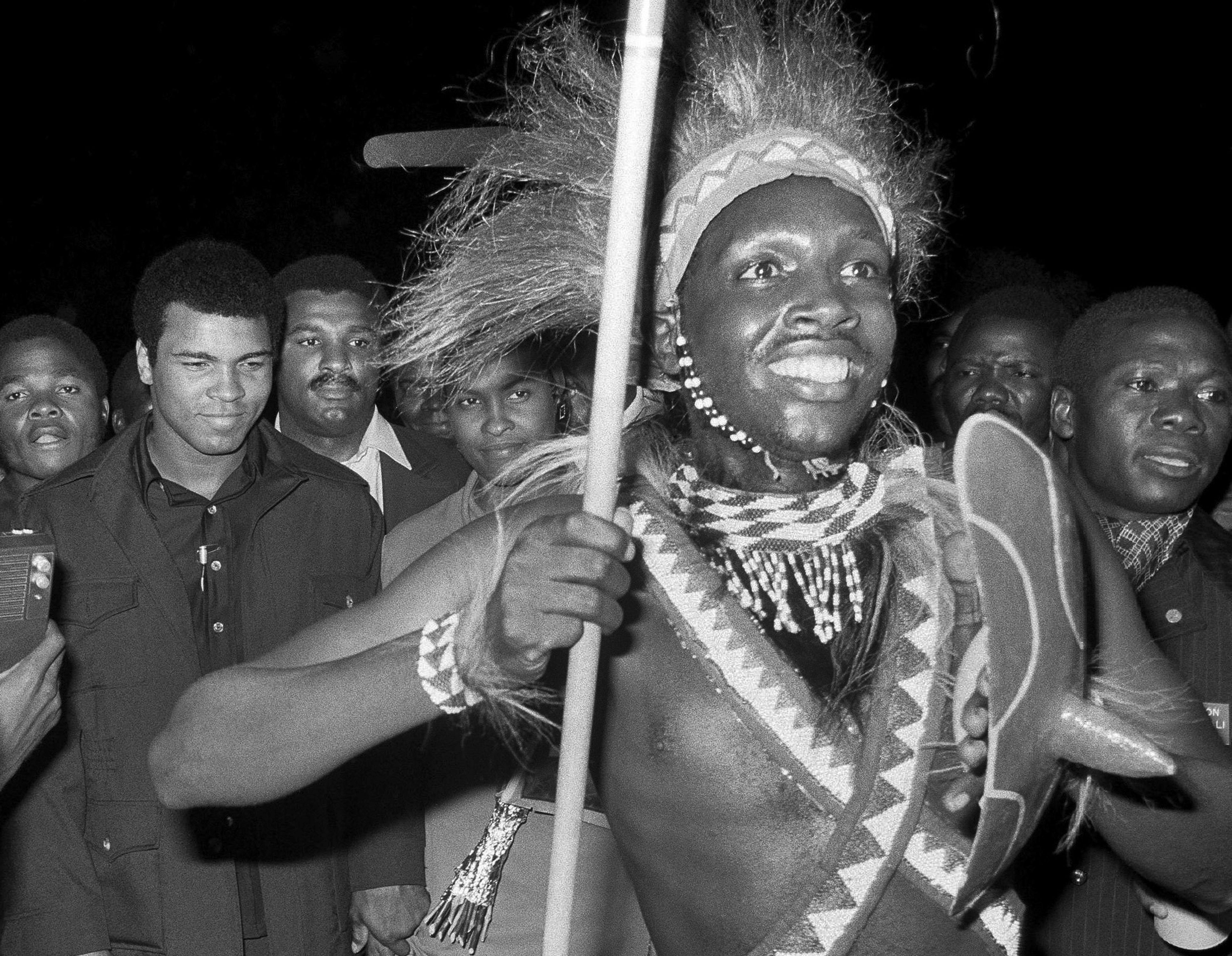 FILE - A Zairian man in traditional clothing carries a spear as he leads Muhammad Ali through a crowd at the airport in Kinshasa, Zaire on Sept. 10, 1974. (AP Photo/Horst Faas, File)