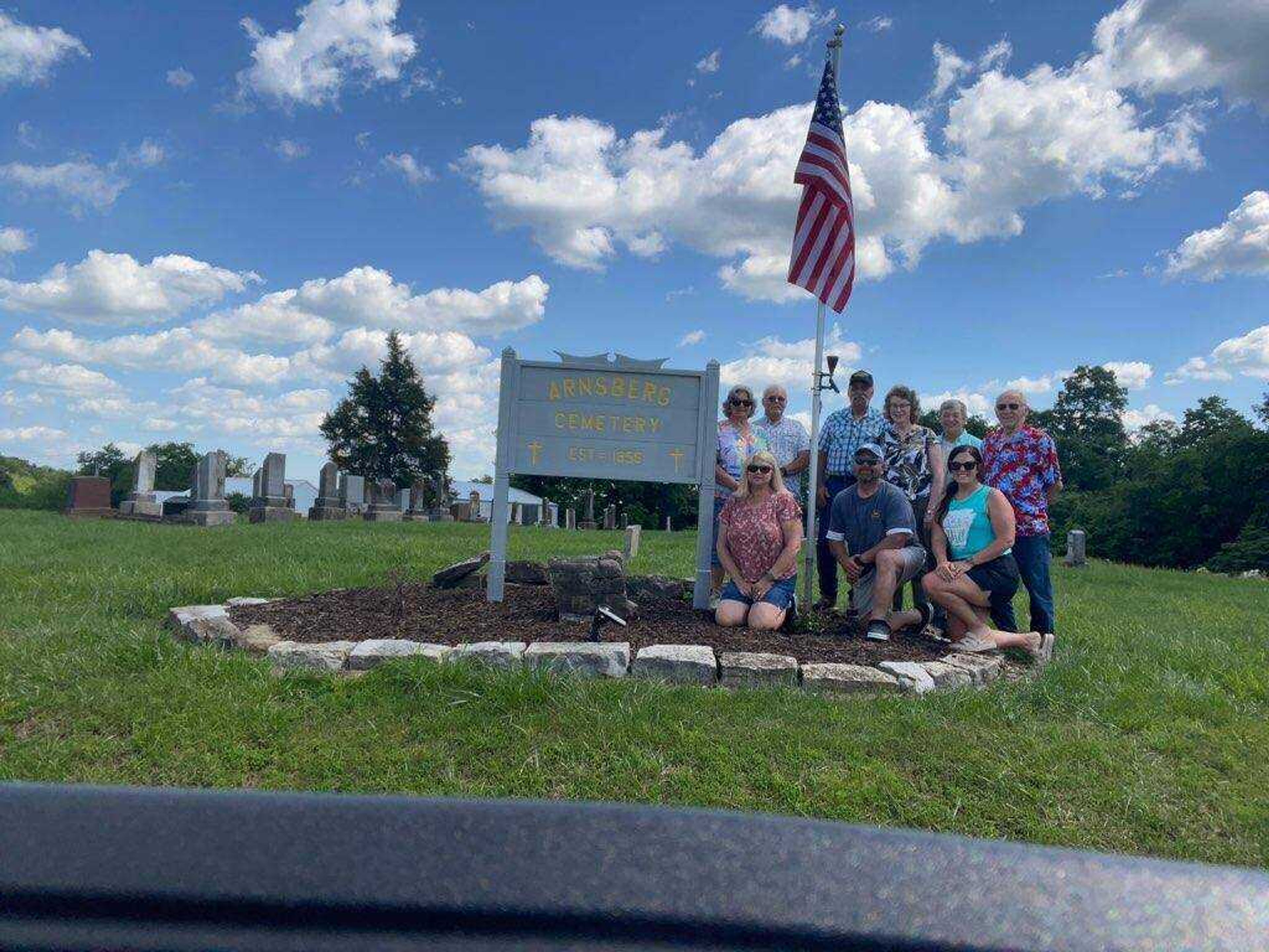 Arnsberg Cemetery Association members gather around the entrance sign and flag pole following the 25th annual business meeting May 19, 2024.  Pictured kneeling, left to right:  Diann Ulmer, Randy Georger, and Rachel Harper; standing, left to right:  Joyce Reed, Mike Reed, Rodney Tucker, Linda Tucker, Sue Sewing, and Dan Dambach.