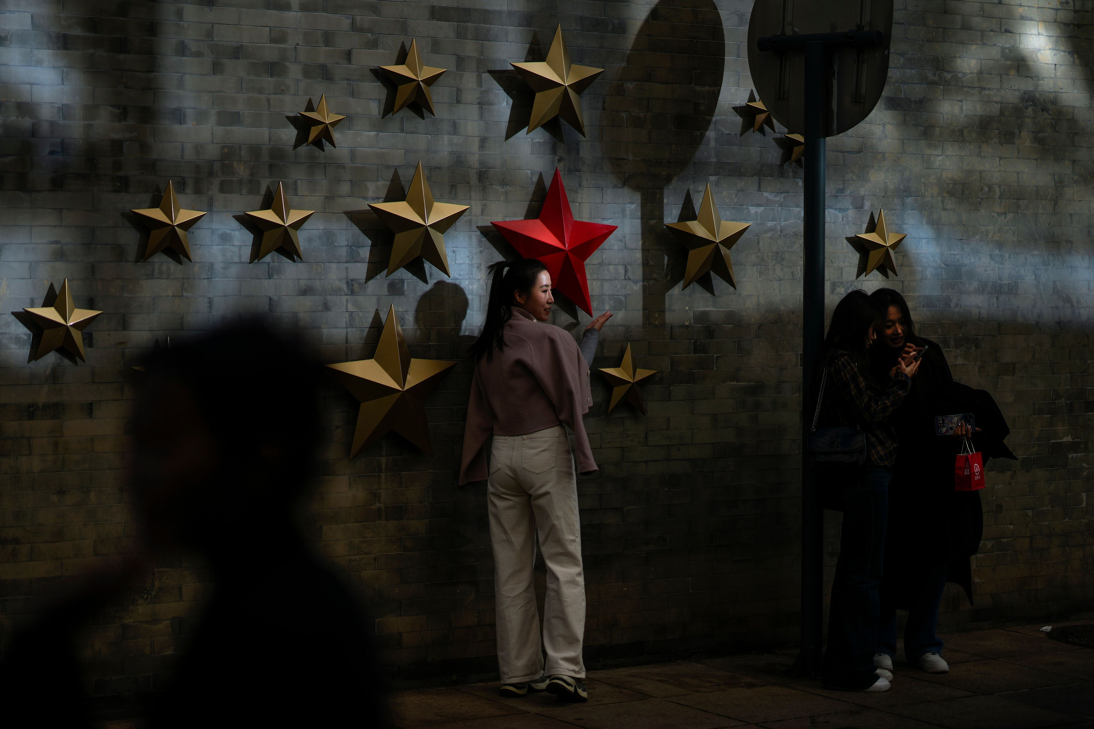 A beam of sunlight is cast on a woman as visitors take souvenir photos with the communist symbol of stars on display along a street in Beijing, Thursday, Nov. 21, 2024. (AP Photo/Andy Wong)