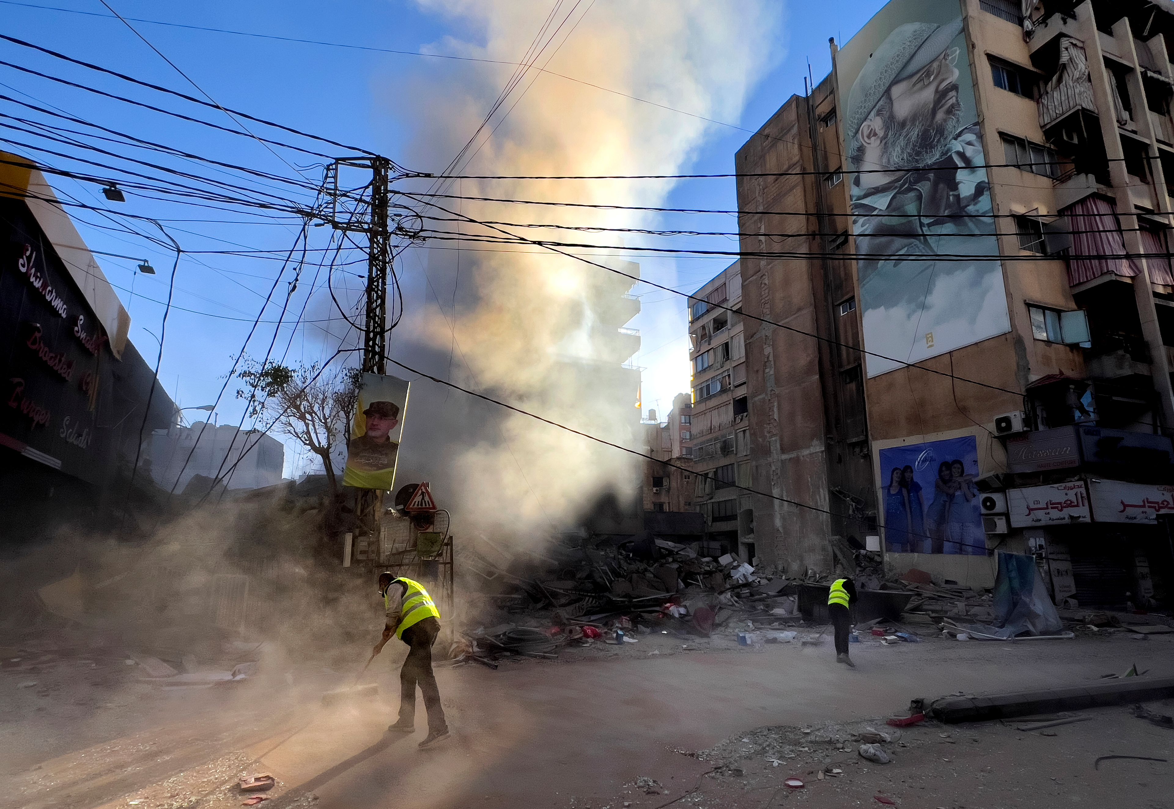 Workers clean a street under a giant portrait of the late Hezbollah military commander Imad Mughniyeh, as smoke rises from a destroyed building that was hit by an Israeli airstrike in Dahiyeh, in the southern suburb of Beirut, Lebanon, Sunday, Oct. 20, 2024. (AP Photo/Hussein Malla)