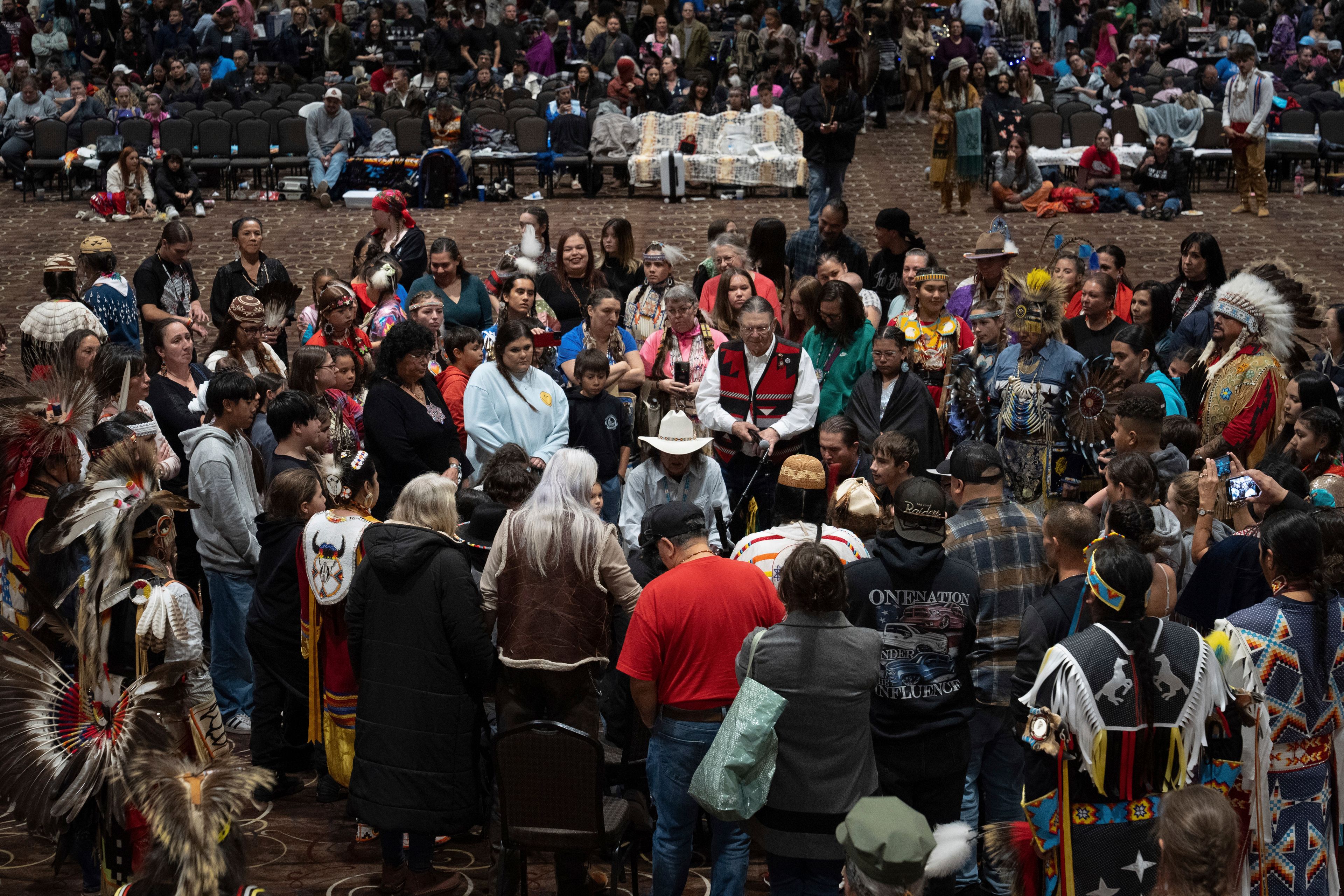 People gather around Chet Clark, in the white hat at center, as he plays an honor song for the Confederated Tribes of Siletz Indians during a powwow at Chinook Winds Casino Resort, Saturday, Nov. 16, 2024, in Lincoln City, Ore. Clark, of the drum group Johonaaii, wrote the song in 2007 and sang it for the first time during the tribe's 30th restoration powwow. It has been sung during every restoration powwow since then. (AP Photo/Jenny Kane)