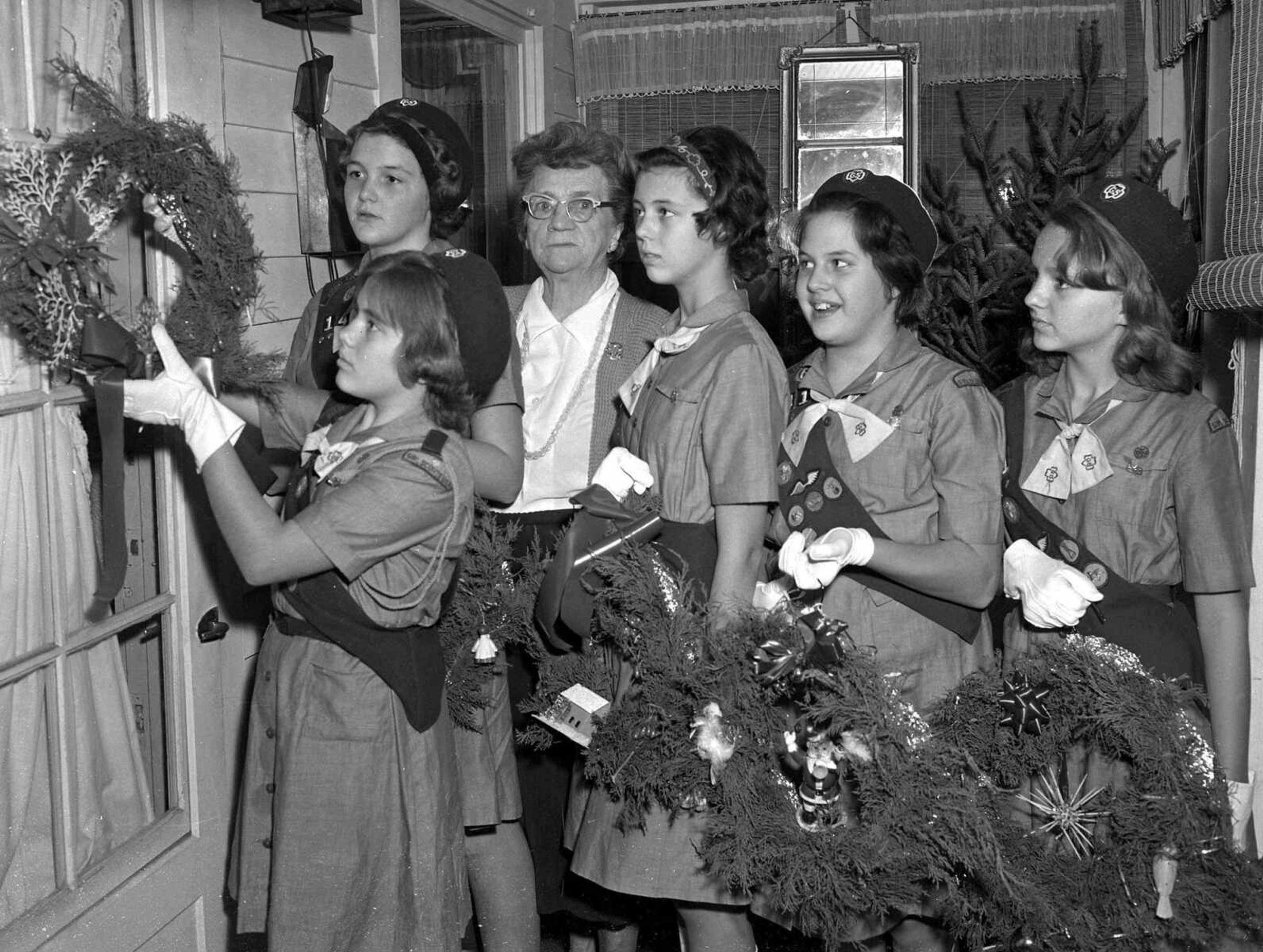 Dec. 21, 1963 Southeast Missourian.
Callers at the city’s nursing homes will note on the doorways this Christmas garlands of cedar gaily ornamented, each differently. Giving of self and service in the spirit of Christmas, Girl Scout Troop 141 of Alma Schrader School cut the cedar, shaped, and placed the ornaments on the wreaths which were delivered this week. Pictured at the Broadway Rest Home, left to right, are Lynda Nations hanging the wreath, Karen Hale, Mrs. Edith Caldwell, owner of the home; Donna  Hitt, Martha Foster and Jane Rodgers. Other members of the troop are: Janet Hedrick, Karen McCall, Judy Fee, Ellen Dye, Dona Kaiser, Deborah Kefauver, Connie Devenport and Jacqueline Kelly. Mrs. Bea Rodgers is the troop leader; Mrs. Bobbie Foster the co-leader. (G.D. Fronabarger photo)