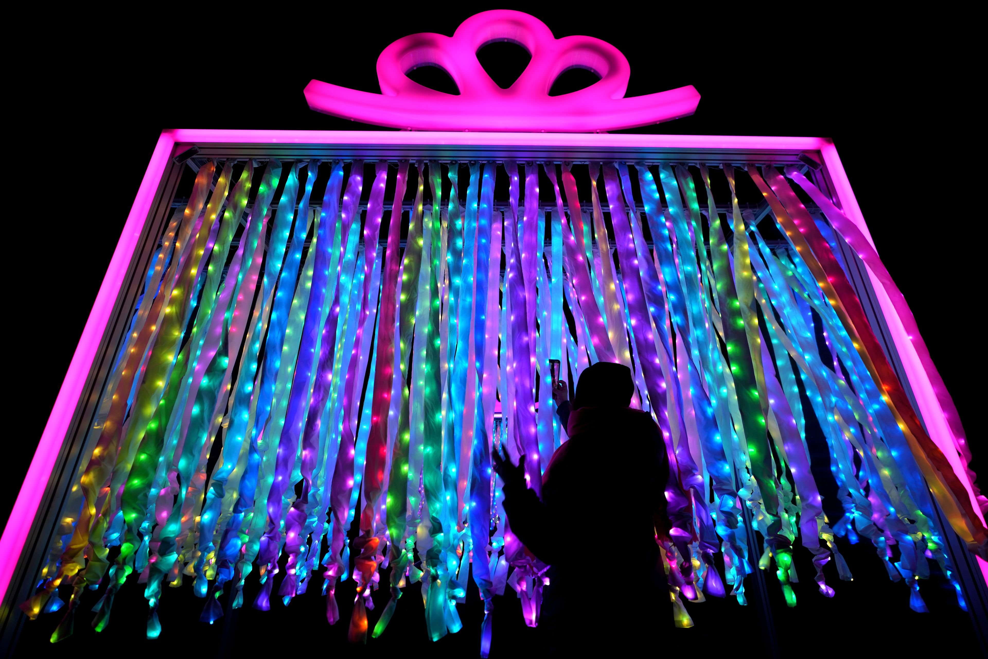 A woman takes a picture at the 'Christmas Presents' on the Christmas light trail as it returns for its 12th year with a showcase of new installations set within the UNESCO World Heritage Site landscape of Kew Gardens in London, England, Tuesday, Nov. 12, 2024. (AP Photo/Kirsty Wigglesworth)