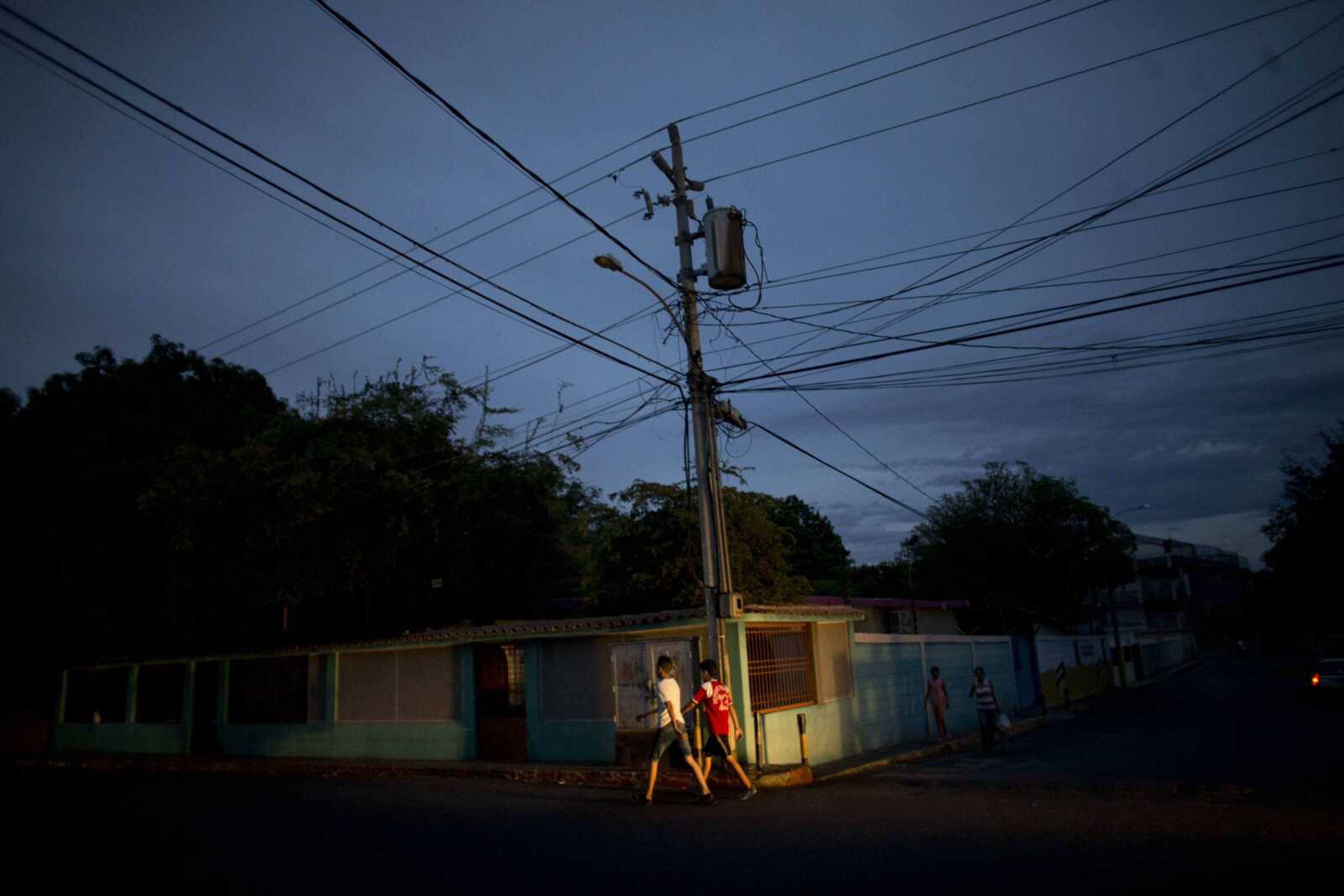 Residents walk under a charred transformer box hanging on a pole that exploded in late March, in La Chinita neighborhood in Maracaibo, Venezuela. Officials repeatedly promised the parts needed to repair it would arrive the next day. So far they haven't come.