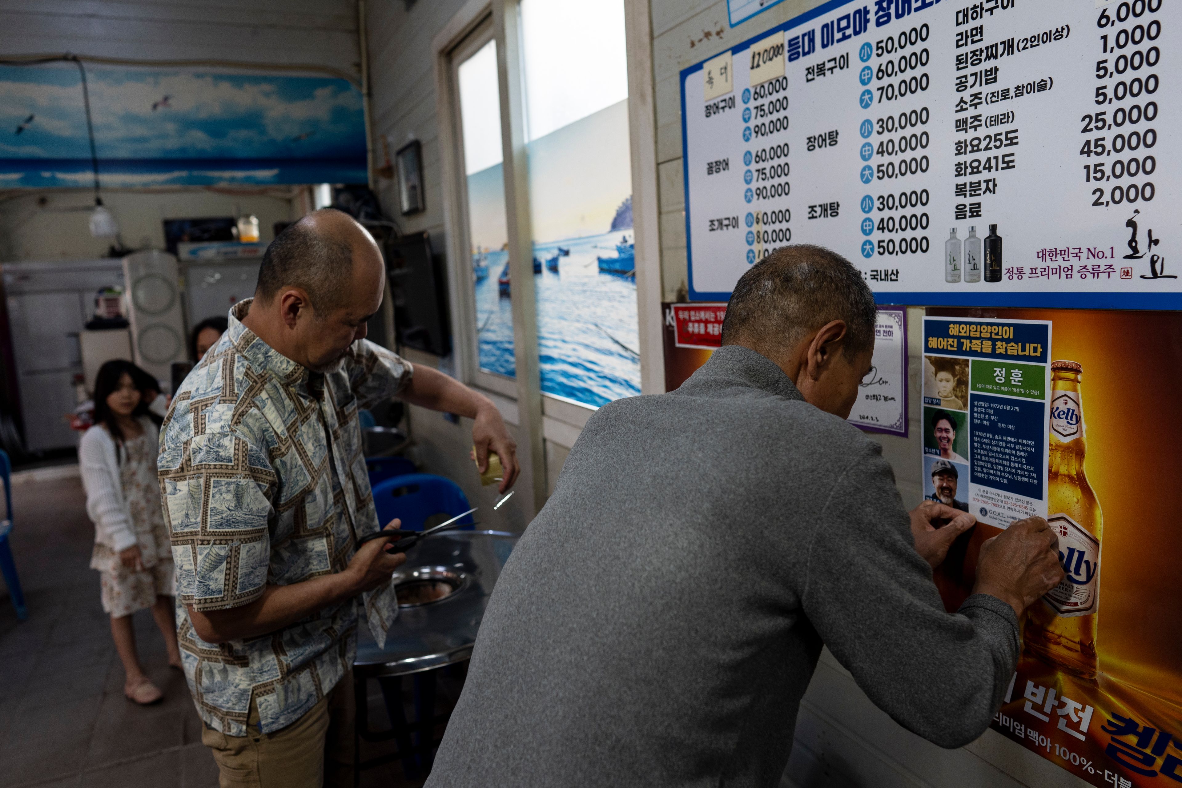 Kenneth Barthel, left, helps Shin Byung-chul post a flyer with photos of Barthel at various ages in his life, on the wall of his restaurant in Busan, South Korea, Friday, May 17, 2024, as Barthel's daughter, Amiya, rear, looks on. Barthel's mother had ordered him soup in a restaurant in the area when he was 6 years old, went to the bathroom and never returned. (AP Photo/Jae C. Hong)