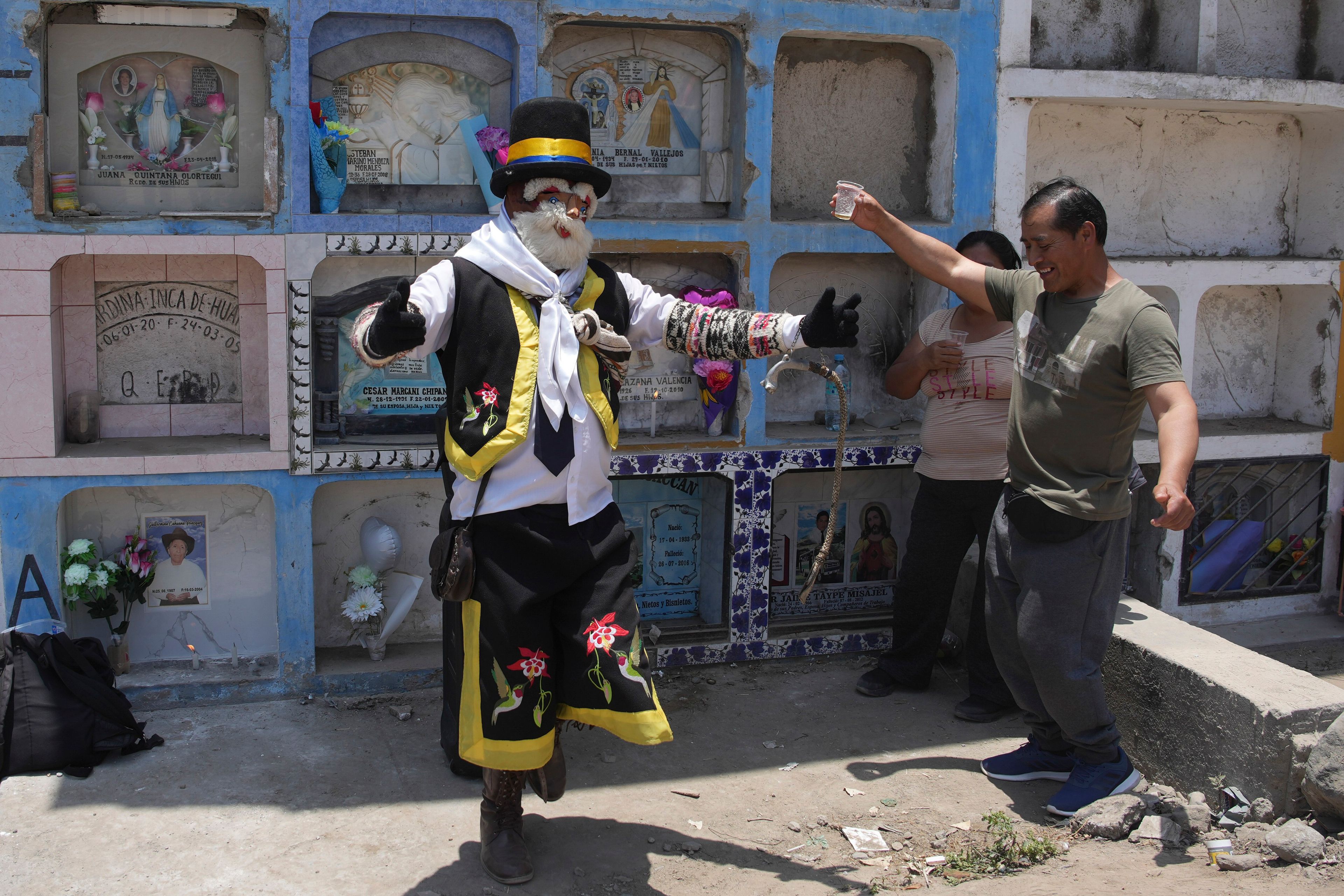 A performer dressed as the Andean character Chuto dances with Oswaldo Bellido who is visiting his parents' grave, during the Day of the Dead celebrations in Lima, Peru, Friday, Nov. 1, 2024, (AP Photo/Guadalupe Pardo)