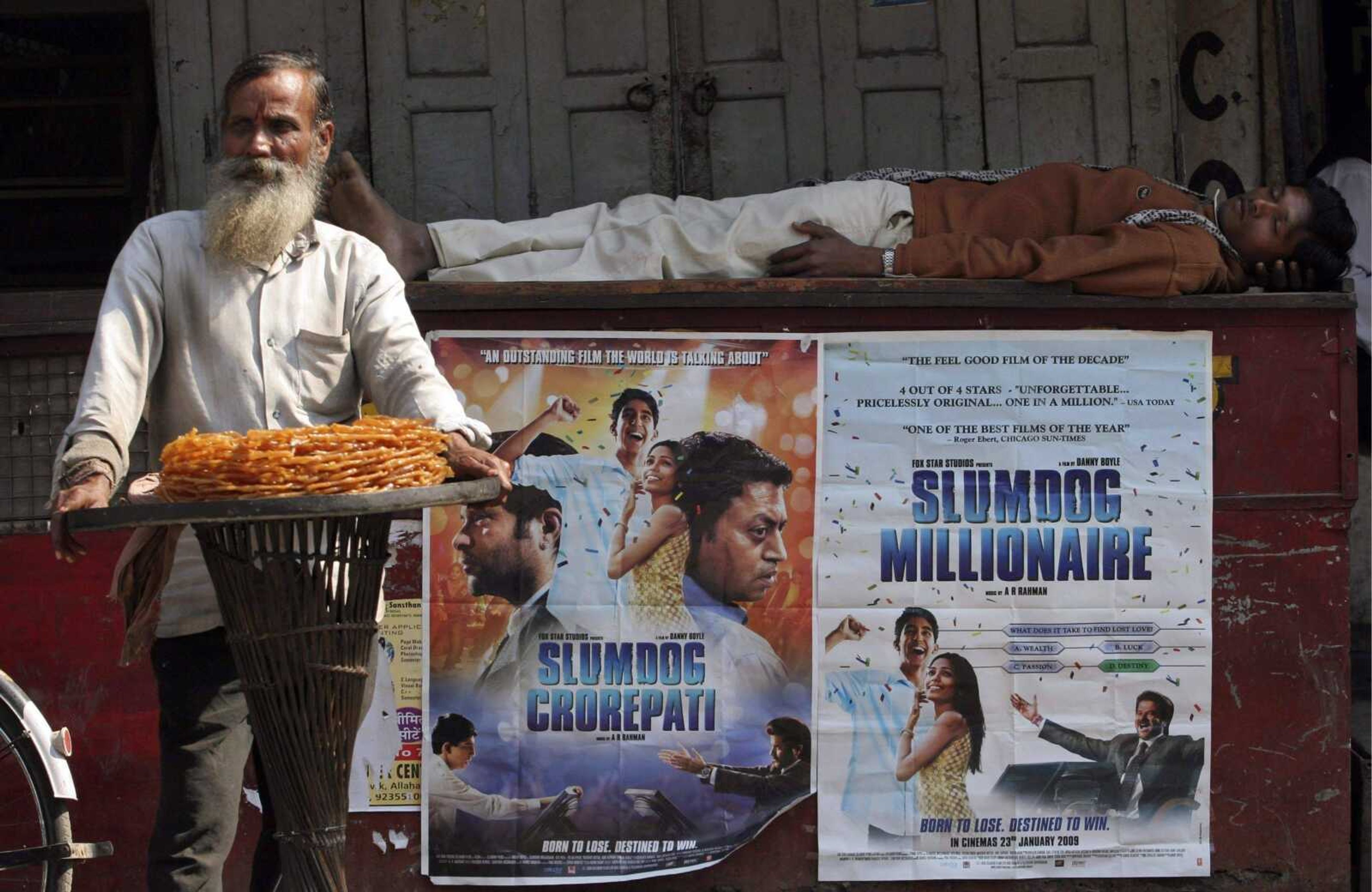 A roadside vendor sells sweetmeats Friday as a man rests on a wall with posters of "Slumdog Millionaire" and its Hindi version, "Slumdog Crorepati." The rags-to-riches story hit Indian theaters Friday to rave reviews and predictions of blockbuster sales. (AP Photo/Rajesh Kumar Singh)